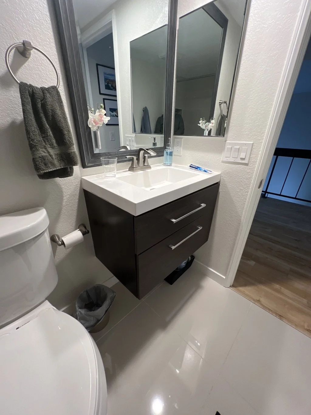 Bathroom with a white sink and dark vanity cabinet, mirror, towel rack with gray towel, toilet, small trash bin, and light switch, with a view into a hallway.