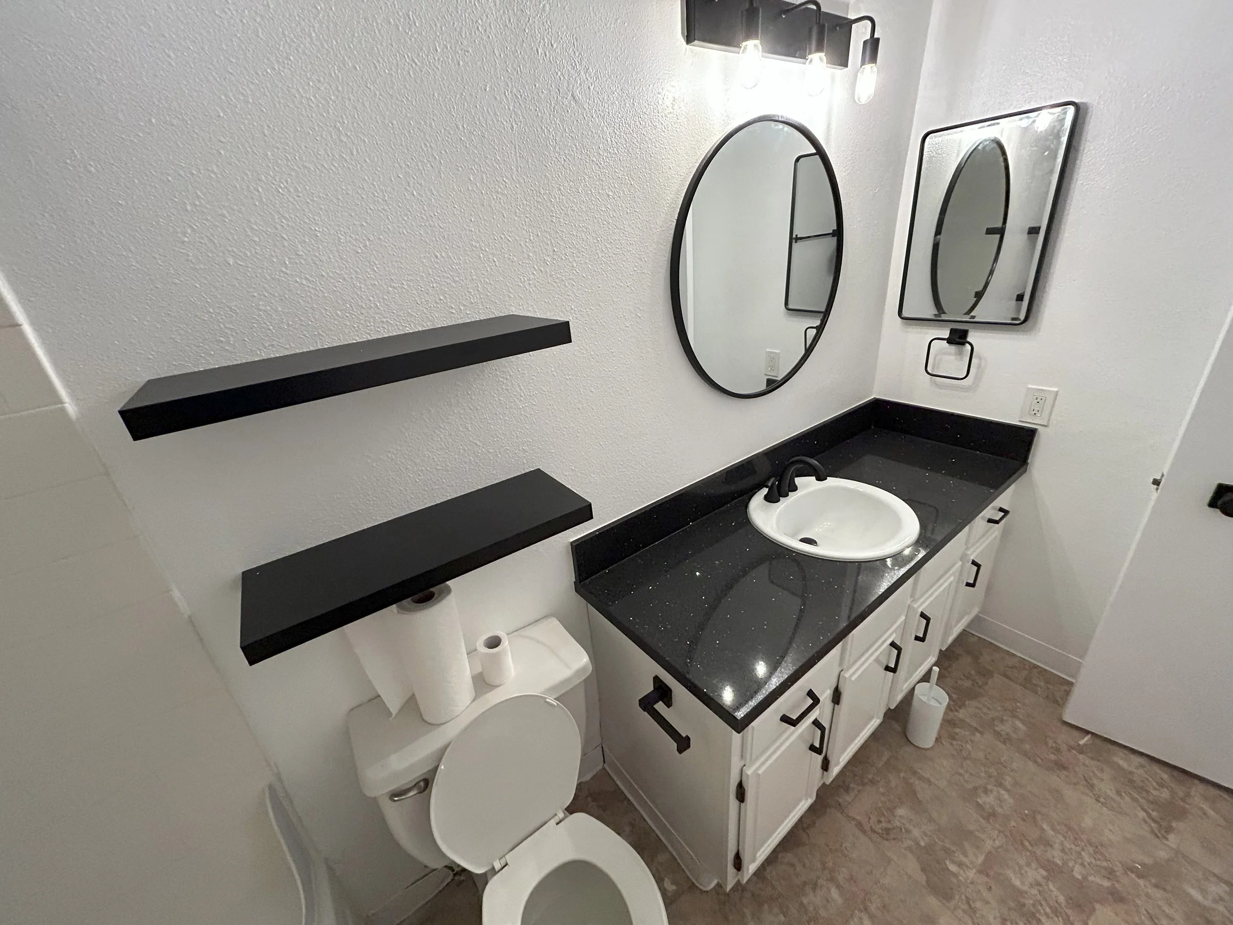 Small bathroom with white walls, black countertop with a white oval sink, two black-framed mirrors on wall, fixtures, toilet with roll of toilet paper, floating black shelves, and beige tile floor.