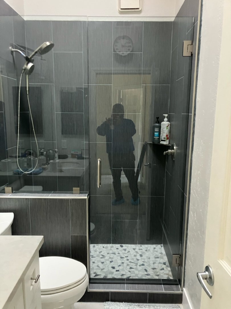 The photo shows a bathroom with a glass shower enclosure, a toilet, and a small sink. Reflecting on the glass, a person is taking the photo, and their reflection is visible.