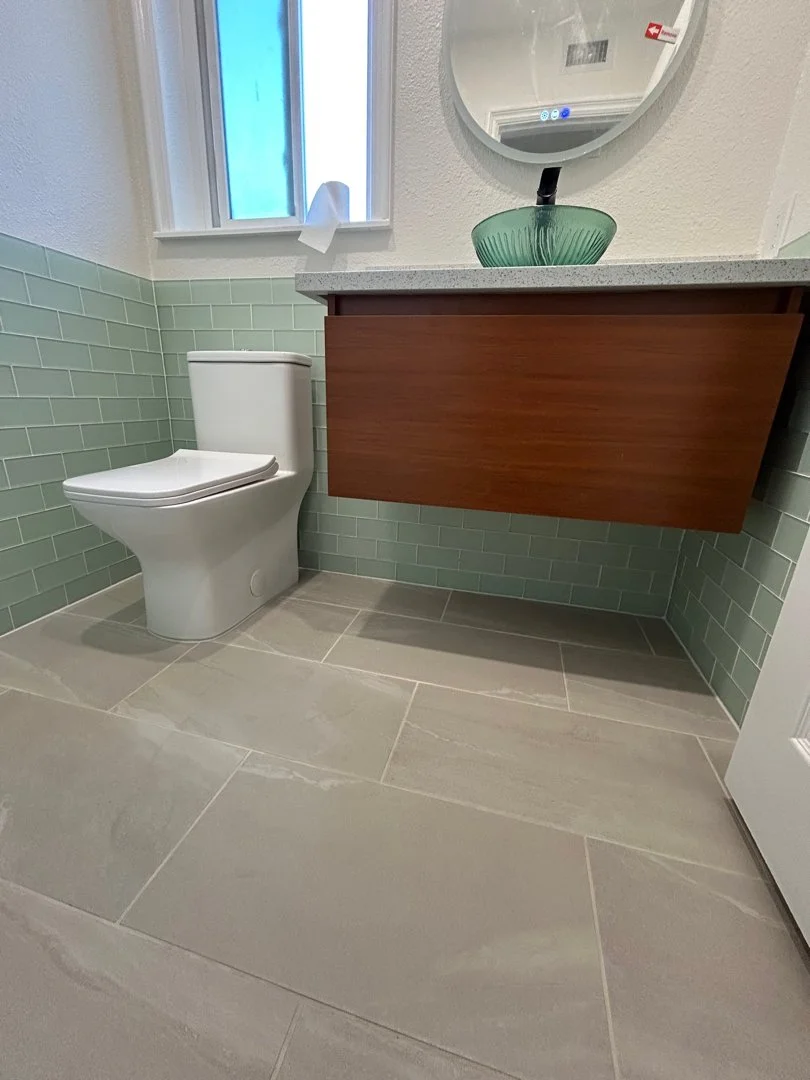 A small bathroom featuring a white toilet next to a wooden vanity with a gray countertop, a green glass vessel sink, a round mirror above the sink, and light gray tile flooring.