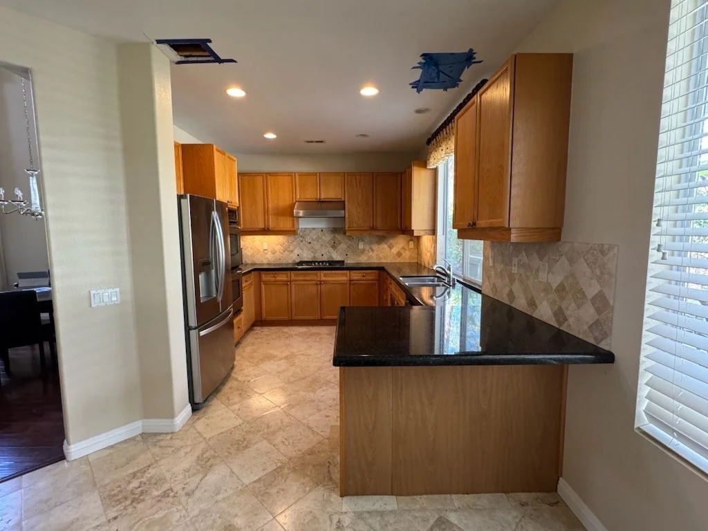 Kitchen with wooden cabinets, black countertop, stainless steel refrigerator, and tiled backsplash, with a window and ceiling lights.