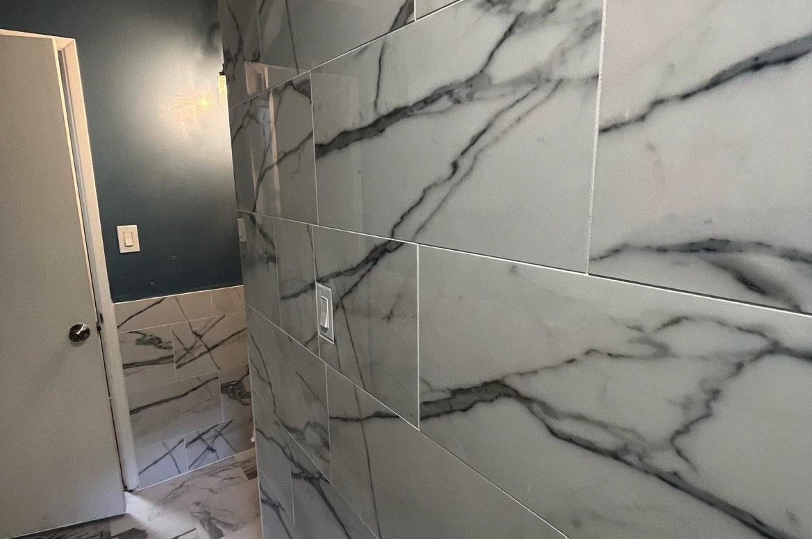 Interior view of a bathroom with white marble wall tiles with dark gray veins, a partially open door, and a wall switch.