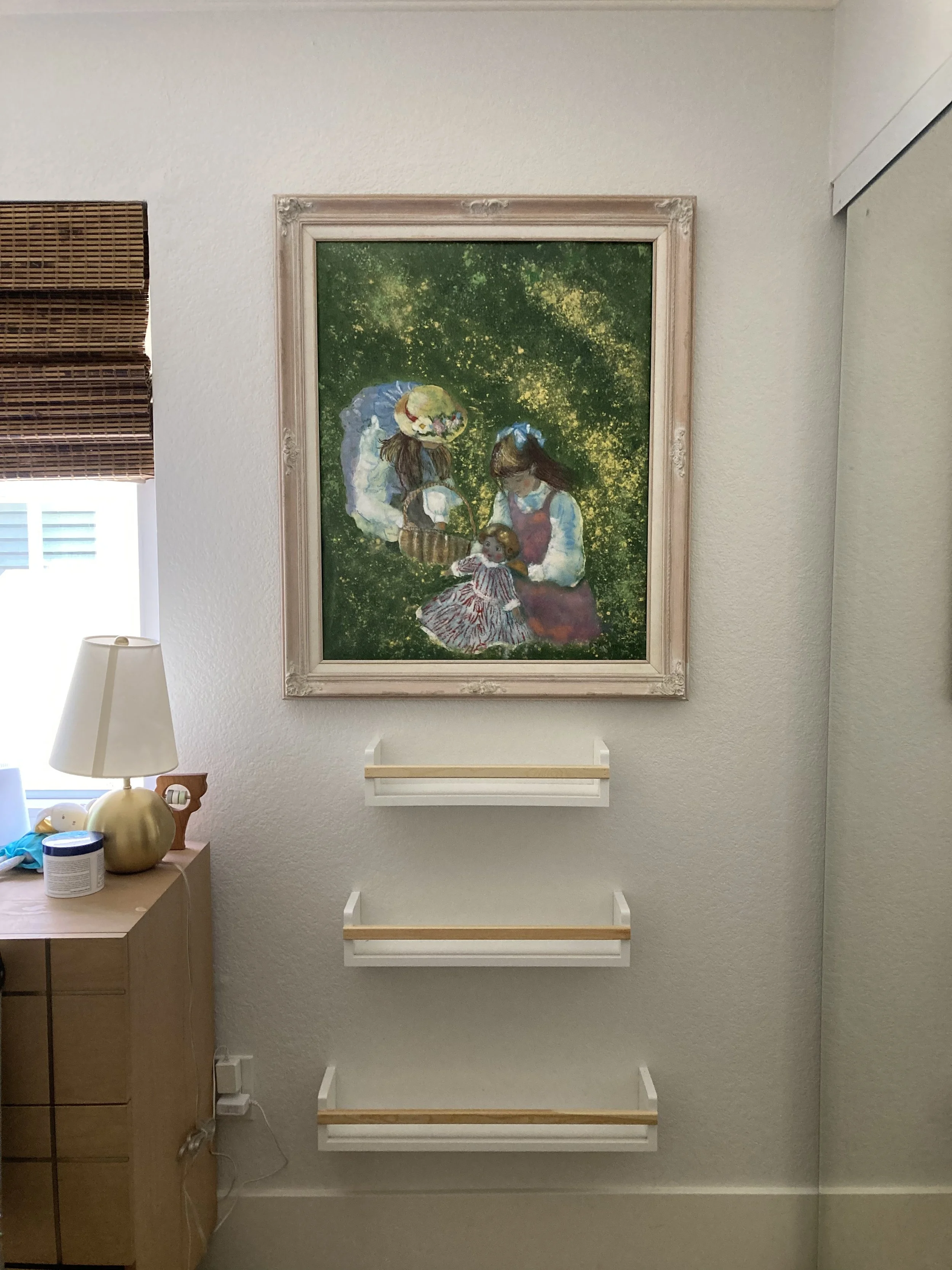 Framed artwork of two women and a child, with the women wearing masks, in green outdoor setting, three white shelves below the picture
