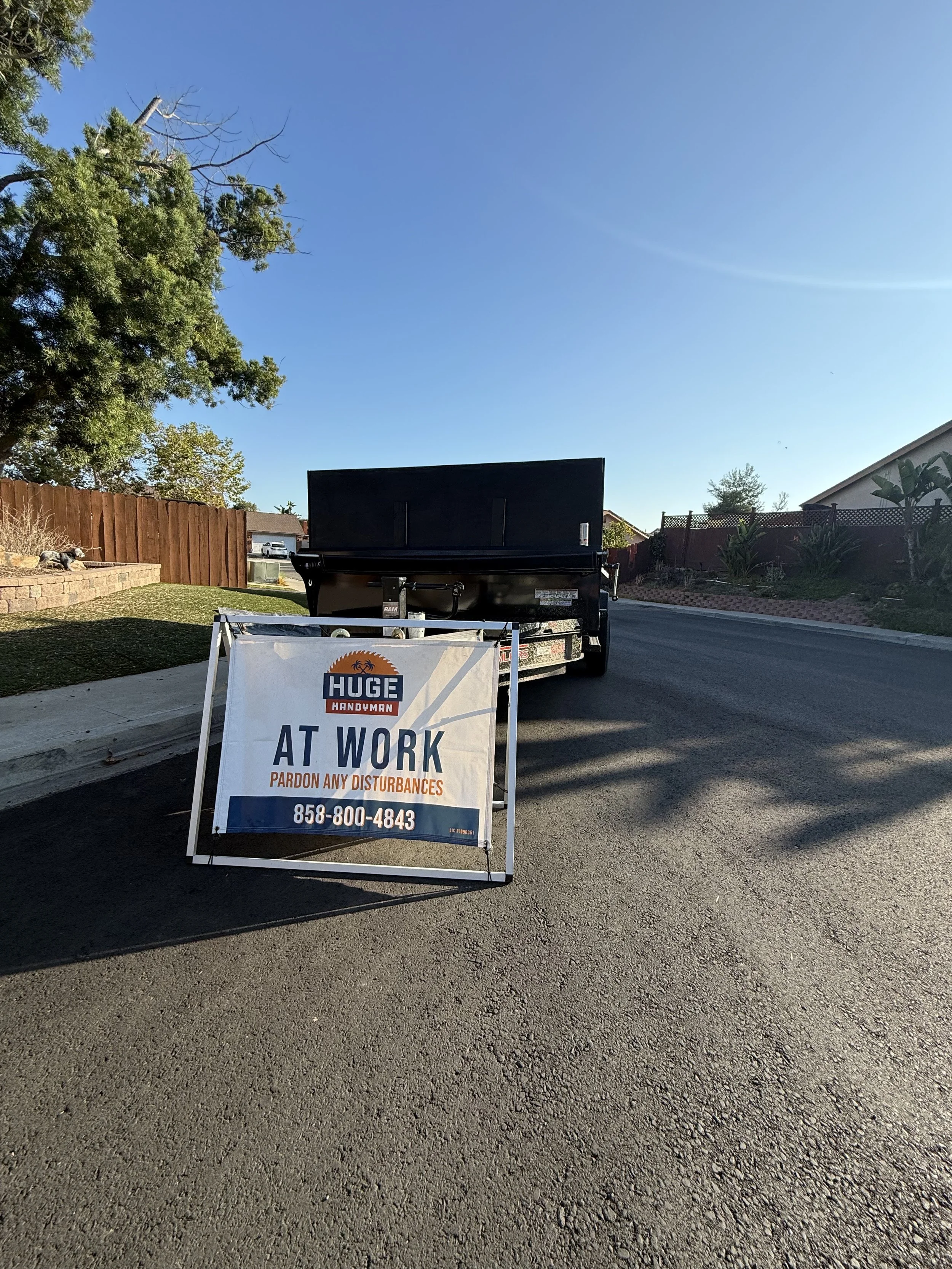 A street scene with a black truck and a sign that reads 'HUGE HANDYMAN AT WORK PARDON ANY DISTURBANCES' along with a phone number, on a sunny day with clear skies.