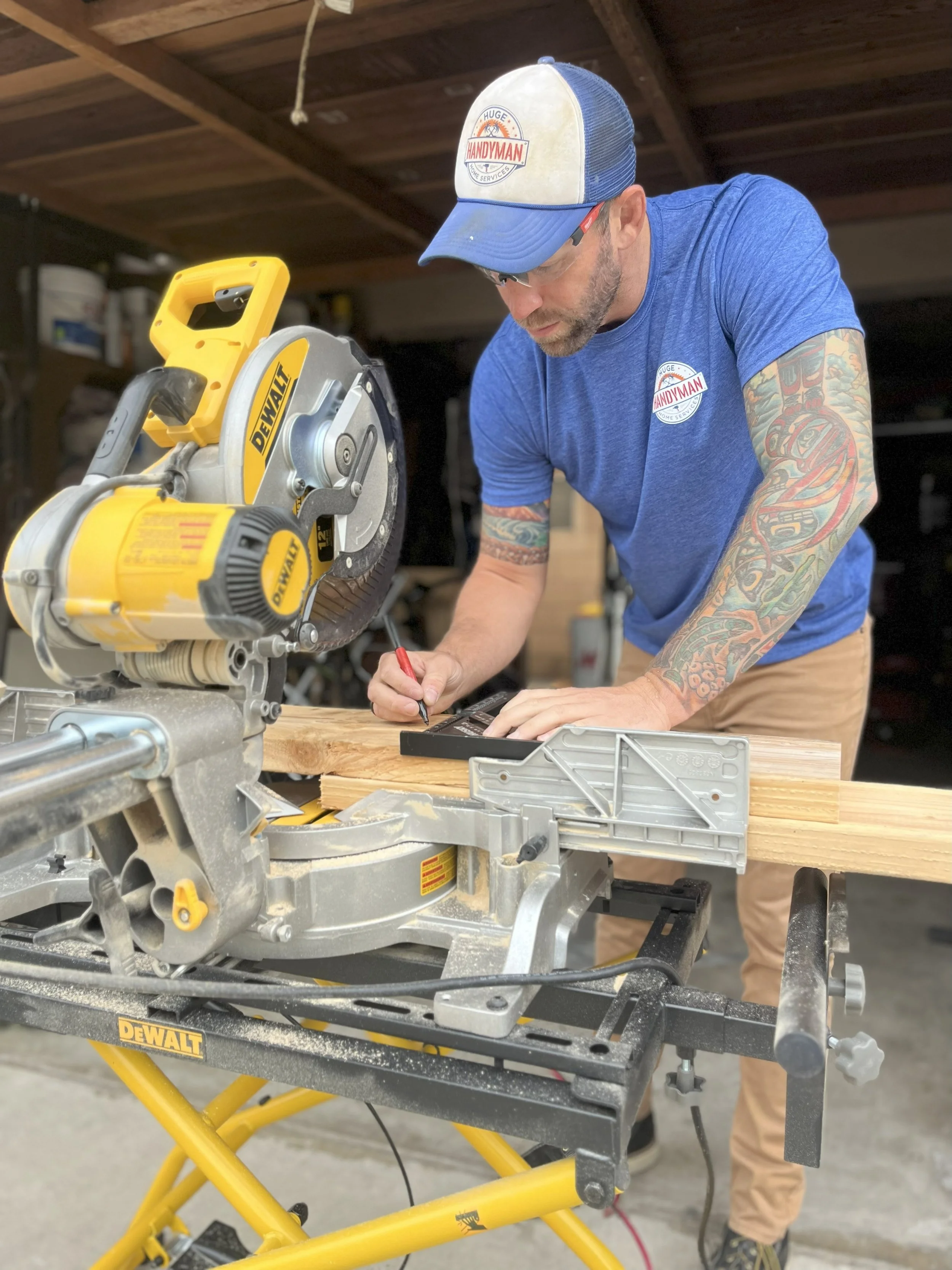 A man with tattoos on his arms is measuring a piece of wood with a square tool while using a yellow DeWalt miter saw in a workshop.