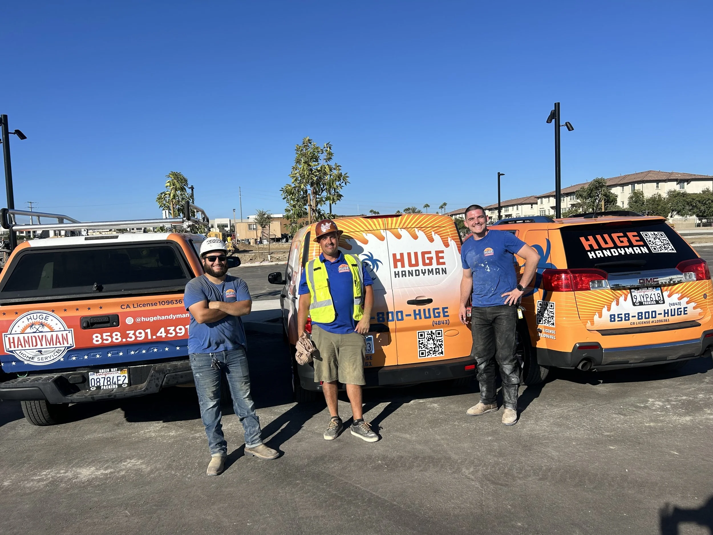 Three men standing in front of branded work vehicles with a bright blue sky background. The vehicles display the logo and contact information for a handyman service called "HUGE HANDYMAN," with orange, blue, and white colors.