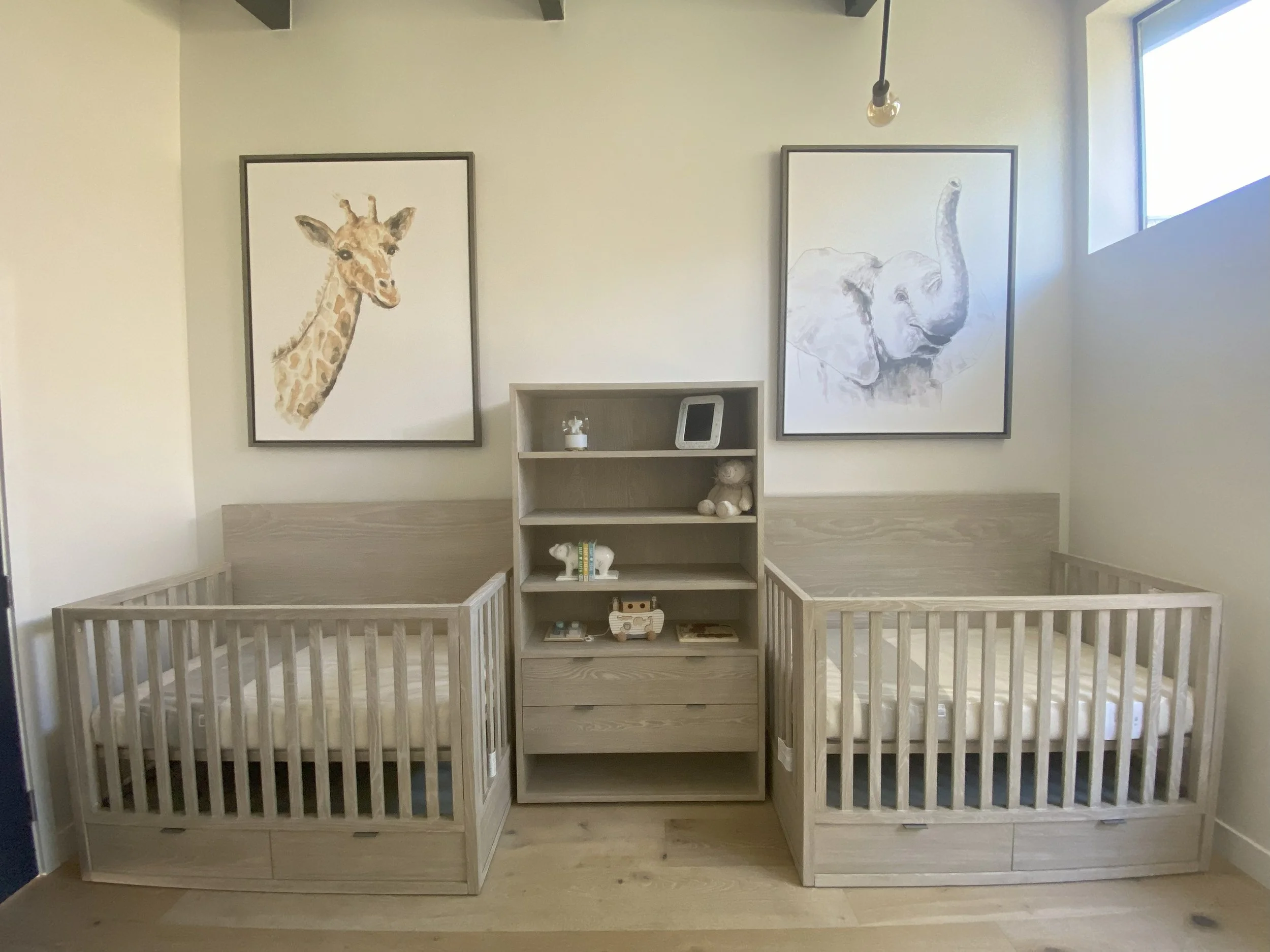 A bedroom with two cribs, one on each side, with a small bookshelf in between. The bookshelf has stuffed animals, books, and decorative items. Above the cribs are two framed animal prints, one of a giraffe and the other of an elephant. There is a window to the right that lets in natural light.
