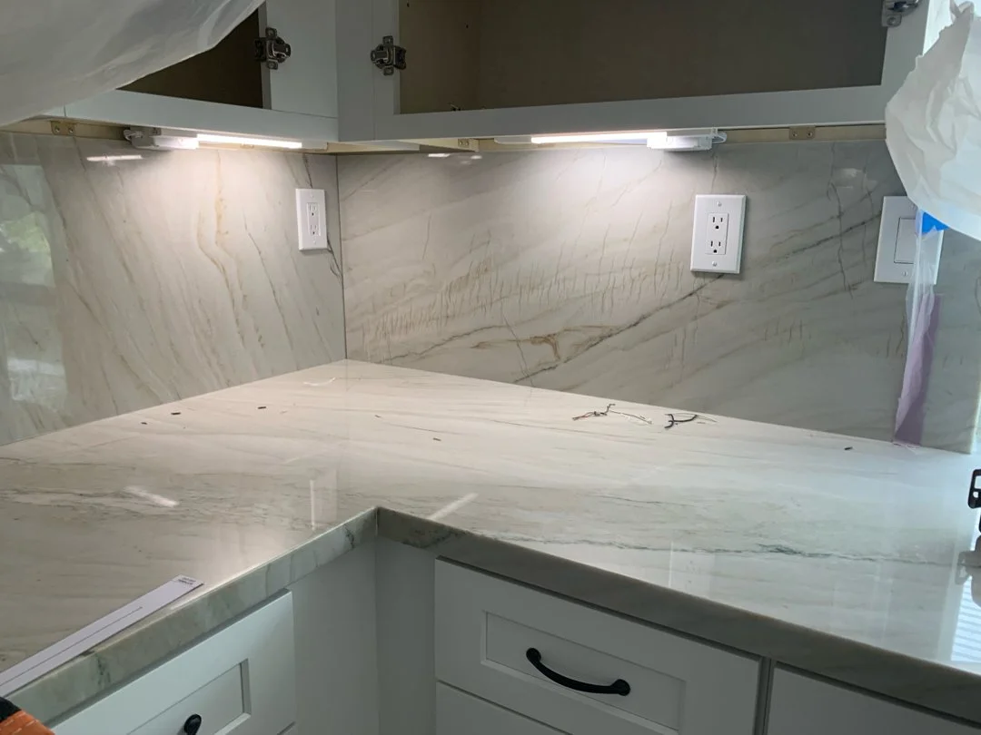 Photo of a kitchen countertop and backsplash made of light-colored marble with gold and gray veins. The countertop has a rounded edge, and there are electrical outlets on the backsplash. Part of an overhead cabinet is also visible.