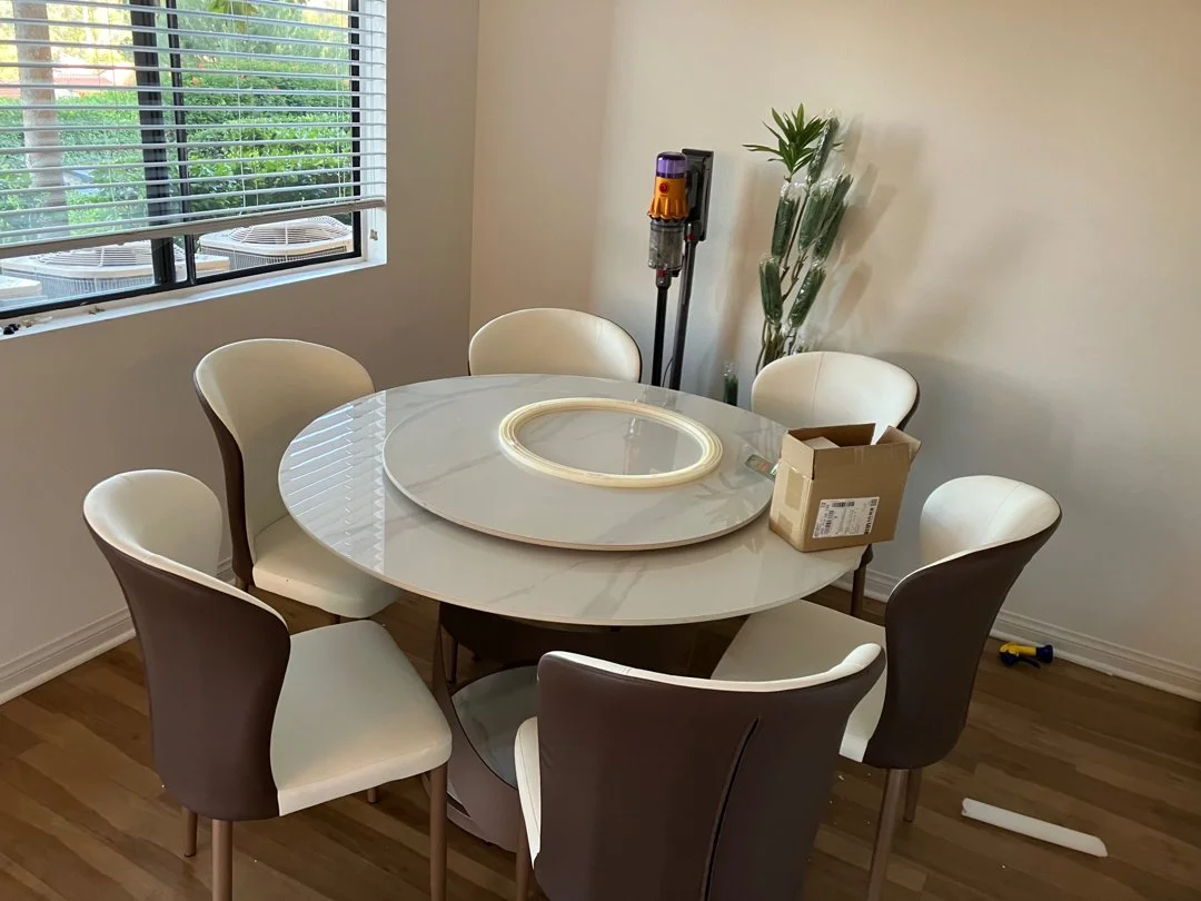 A dining room with six chairs around a round marble table, which has a circular LED light fixture on top. There is a cardboard box and a wall-mounted vacuum cleaner in the corner near a window with blinds.