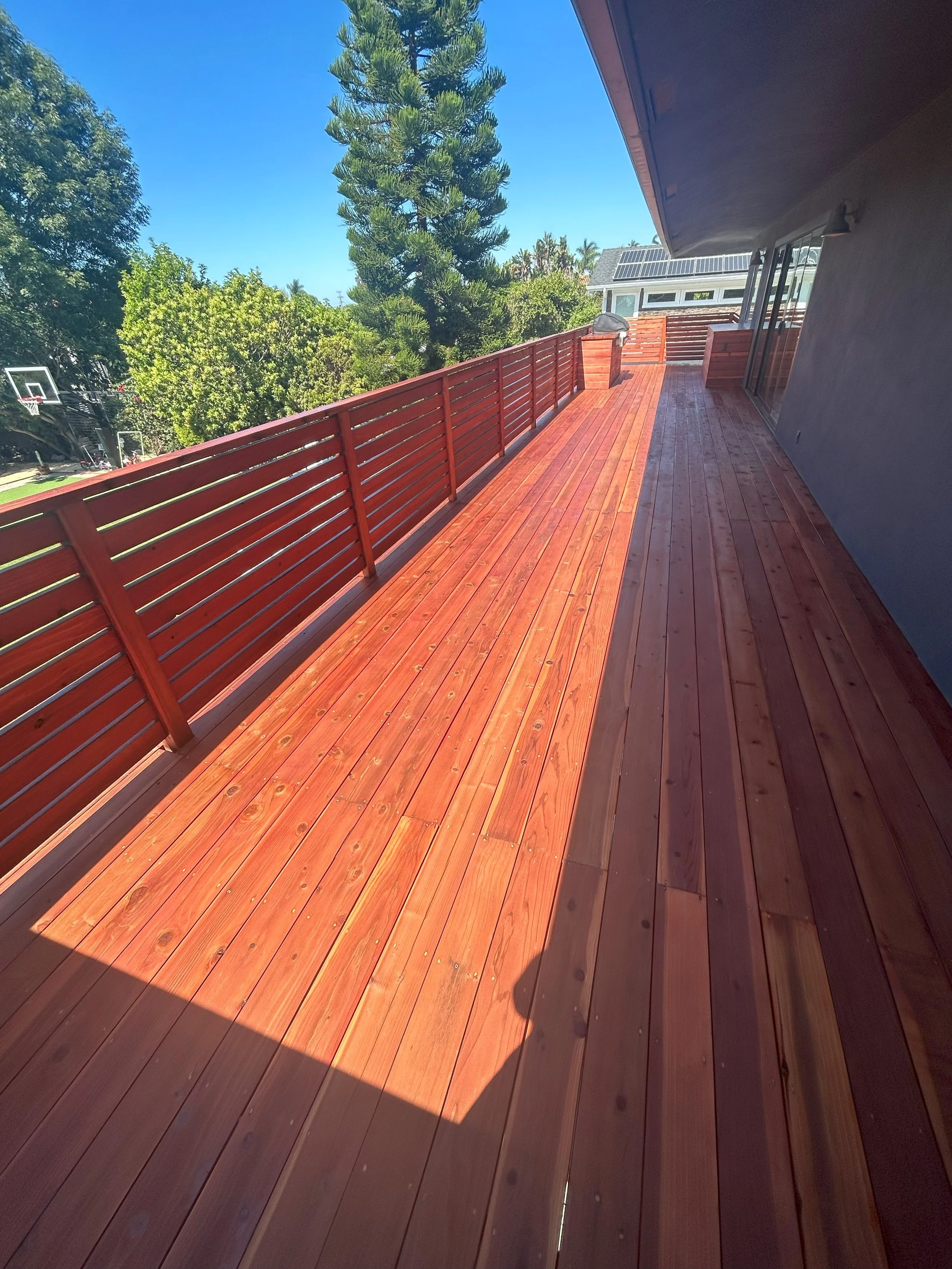A spacious wooden deck with red railing, attached to a house with a glass sliding door. The deck is on an upper level, overlooking a yard with trees and a basketball hoop, under a clear blue sky.