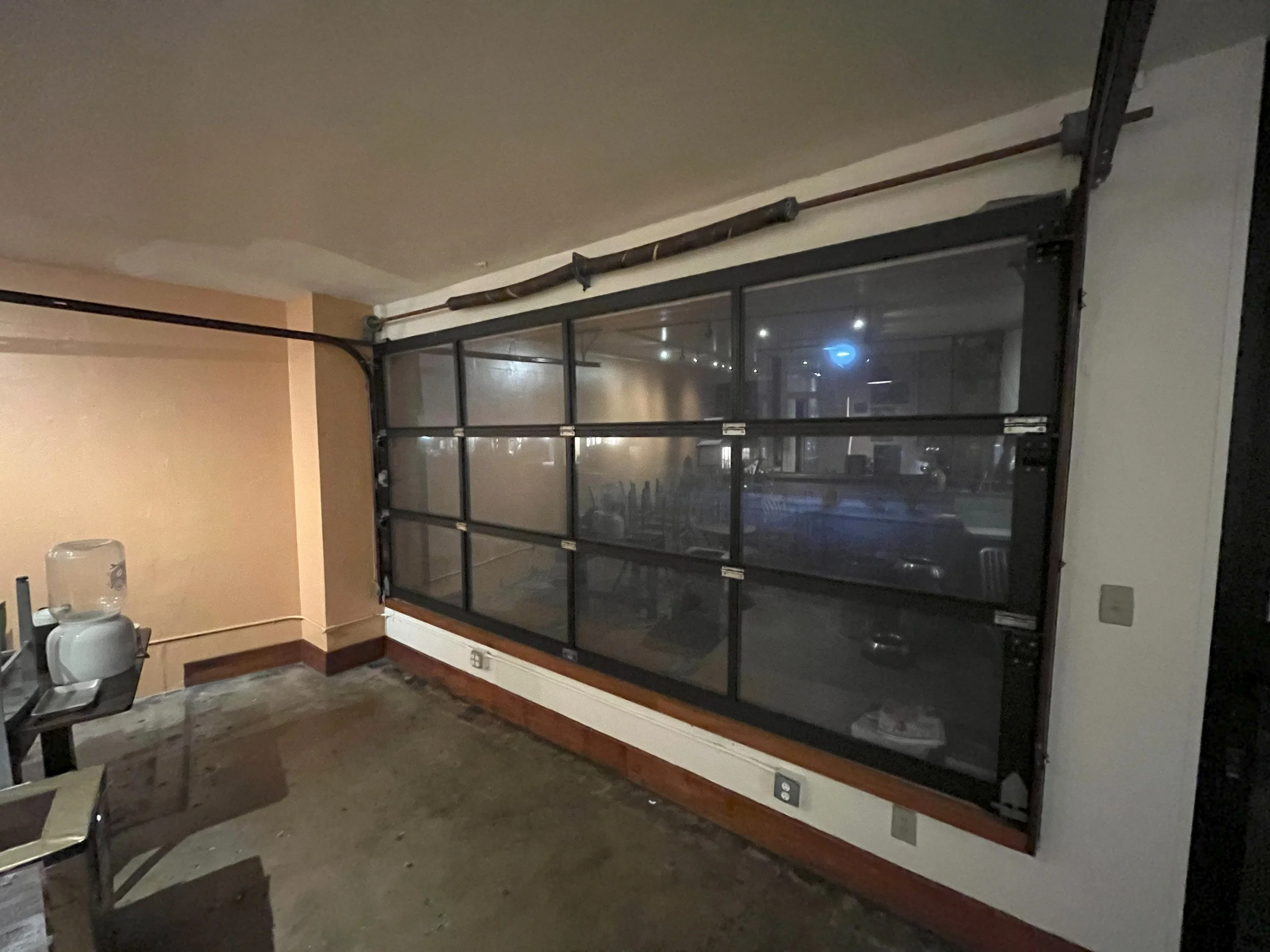 Interior of a room with a large glass garage door, a water dispenser, a chair, and visible electrical outlets, with a concrete floor and beige and white walls.