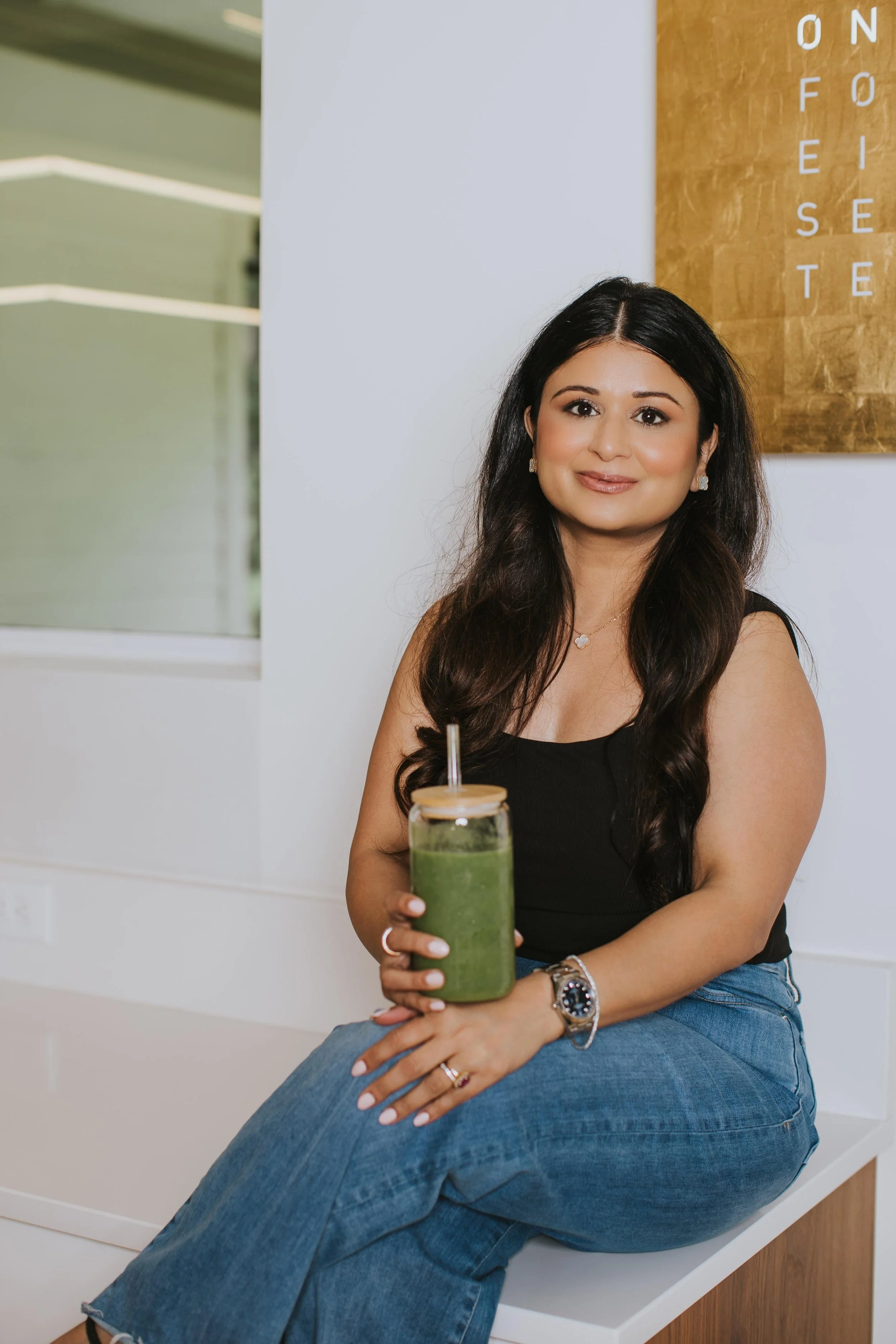 Dr. Sharma smiling while sitting on a counter, holding a green smoothie