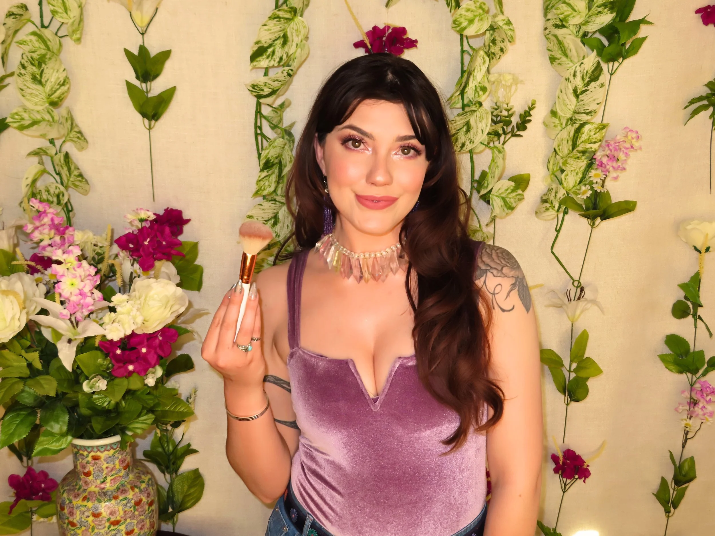 A young woman with long brown hair and tattoos on her shoulder, holding a makeup brush, standing in front of a floral backdrop with pink, white, and green flowers and leaves.