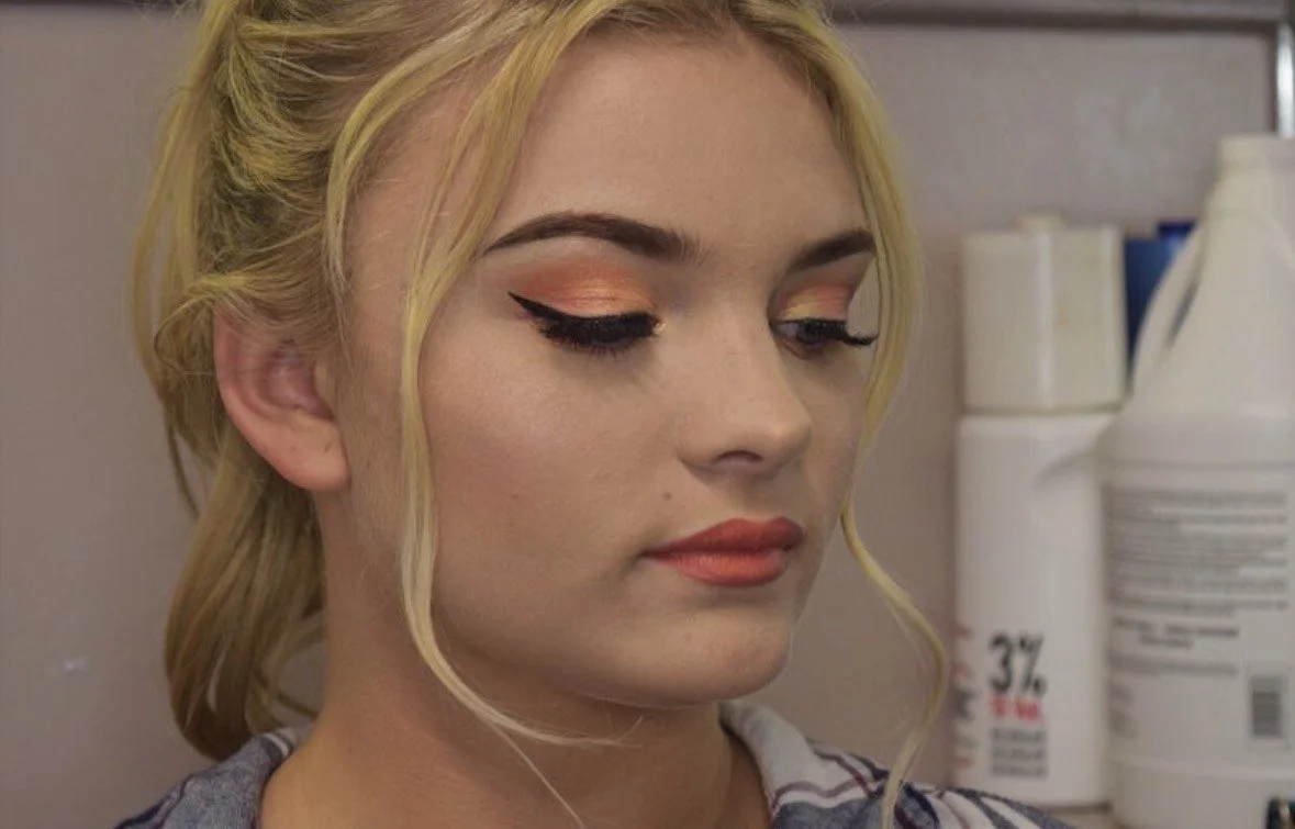 A young woman with blonde hair and makeup with peach eyeshadow and black winged eyeliner, looking down.