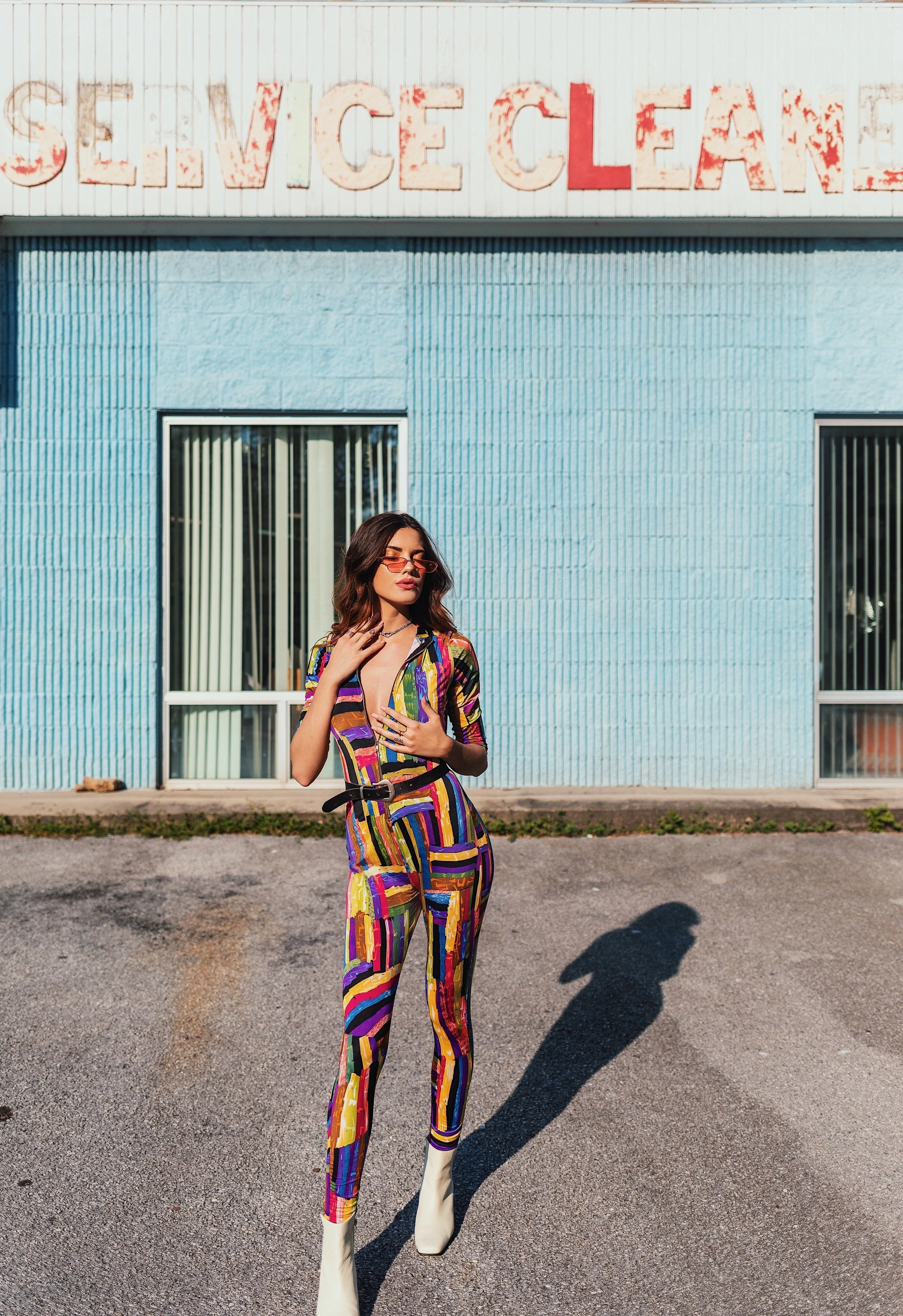 A woman in a colorful, abstract patterned jumpsuit with white boots standing outside in front of a light blue building with a faded sign that reads 'SERVICE CLEAN'.