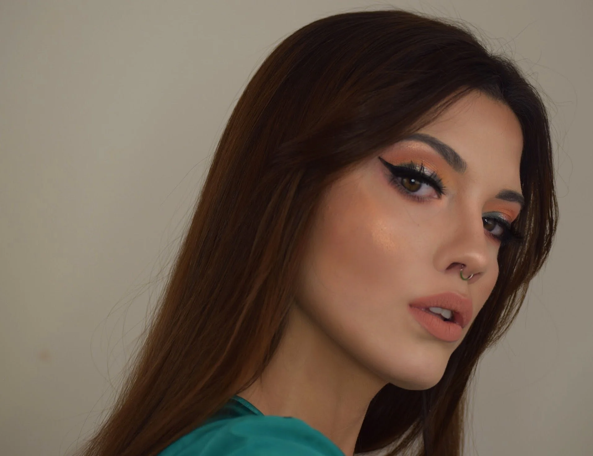 Close-up of a woman with long brown hair, makeup including winged eyeliner and eyeshadow, and a septum piercing, wearing a teal top, against a neutral background.