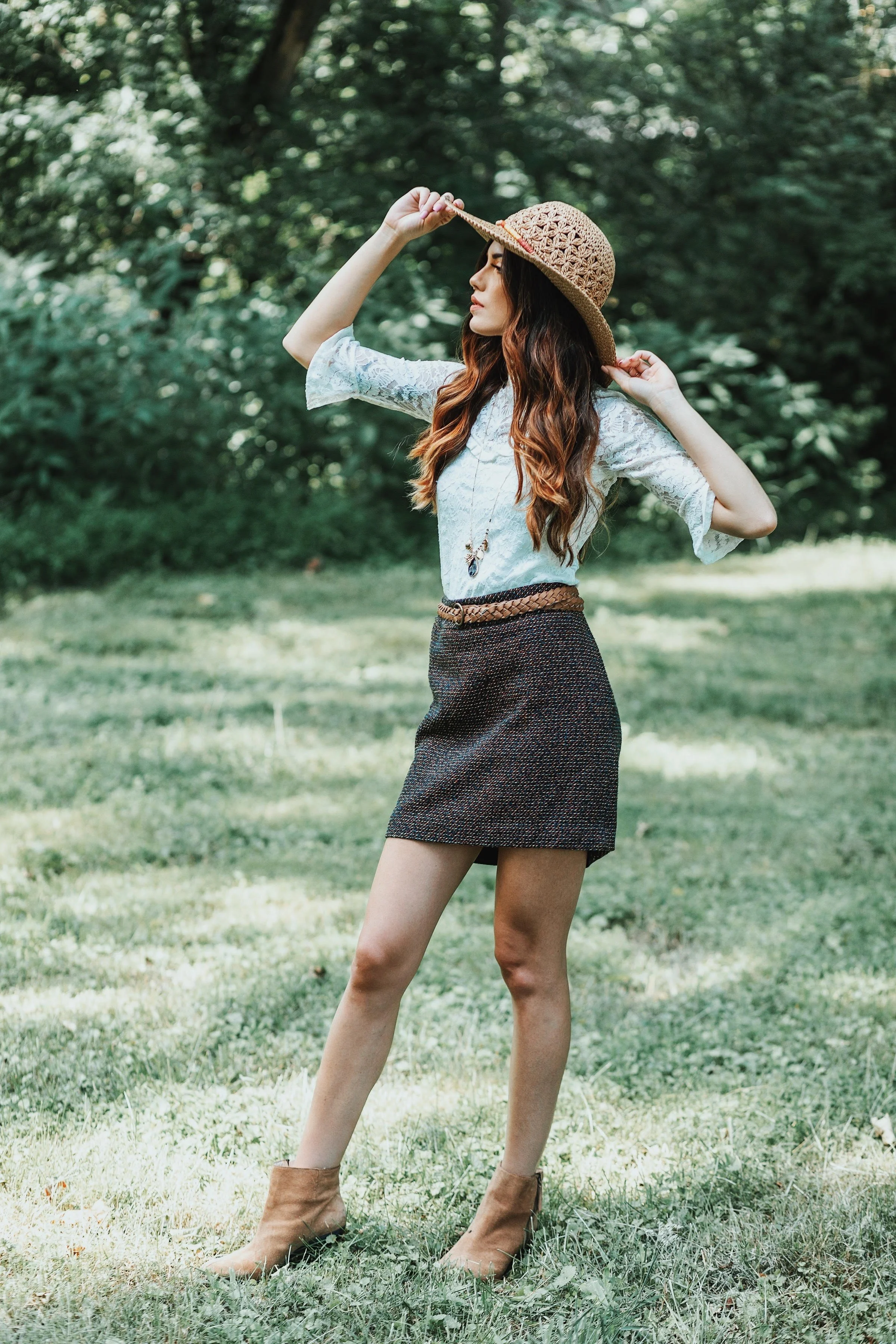 A woman is standing outdoors in a grassy area with trees in the background, wearing a wide-brimmed straw hat, a light blue lace top, a dark skirt, and ankle boots, holding her hat with one hand.