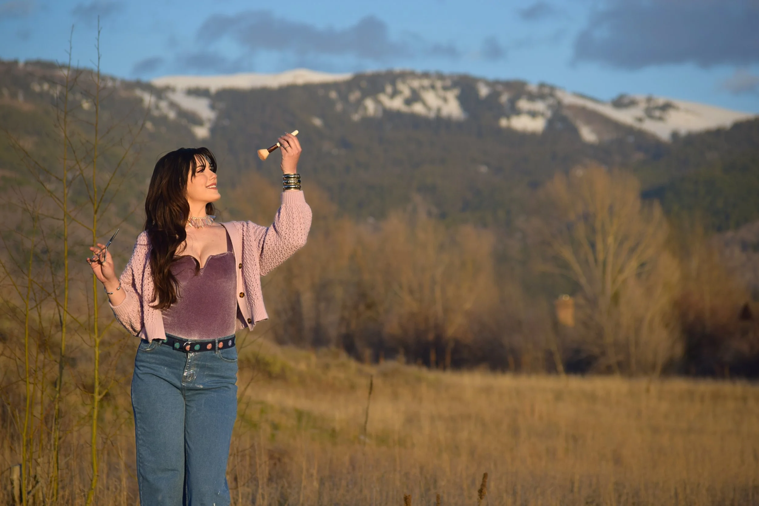 Woman standing outdoors holding a paintbrush and palette, with mountains and trees in the background during sunset or late afternoon.