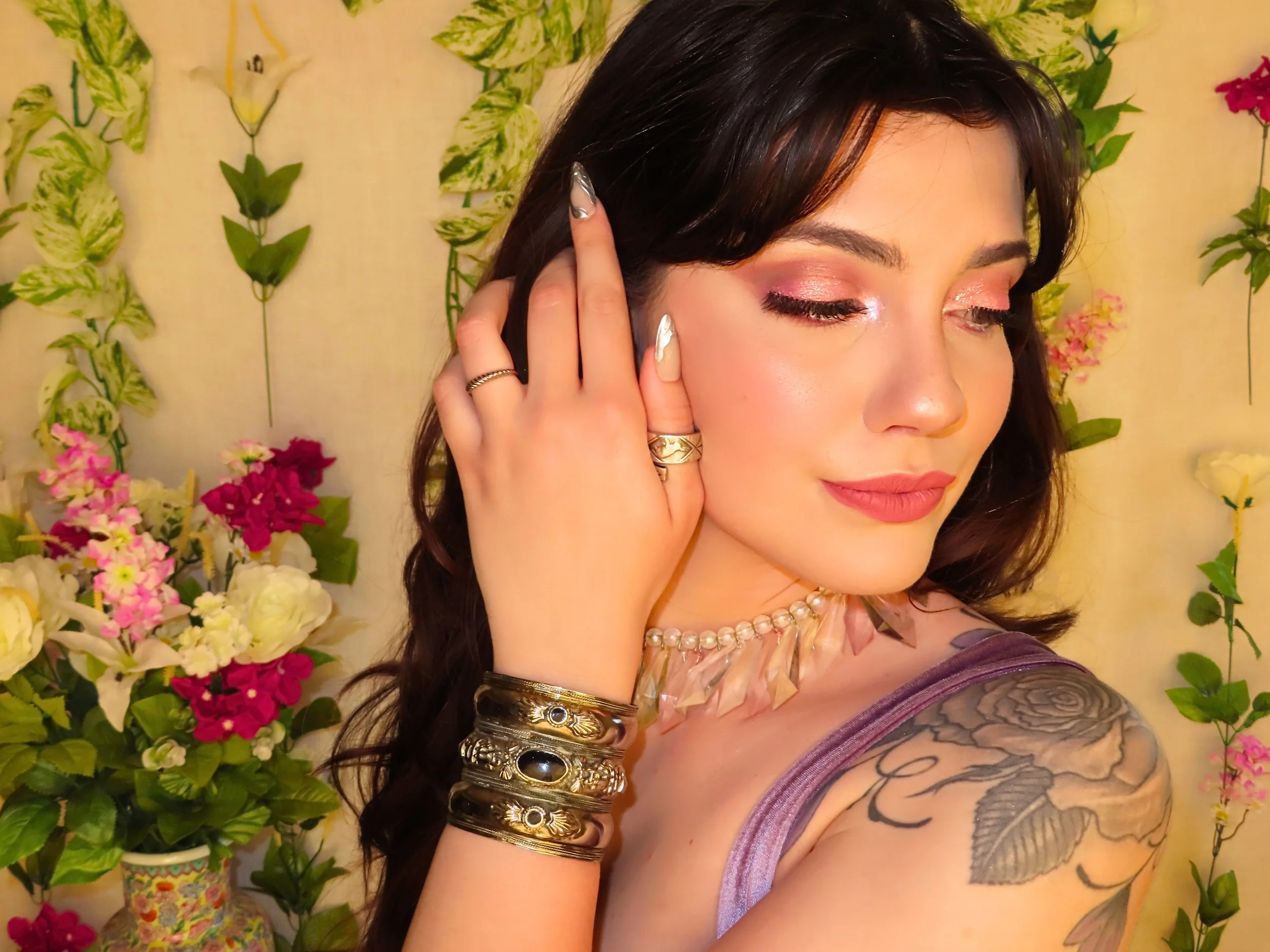 A woman with dark hair and tattoos on her shoulder, wearing makeup, jewelry including bracelets, rings, and a necklace, posing with her hand near her face against a background of various colorful flowers and green leaves.