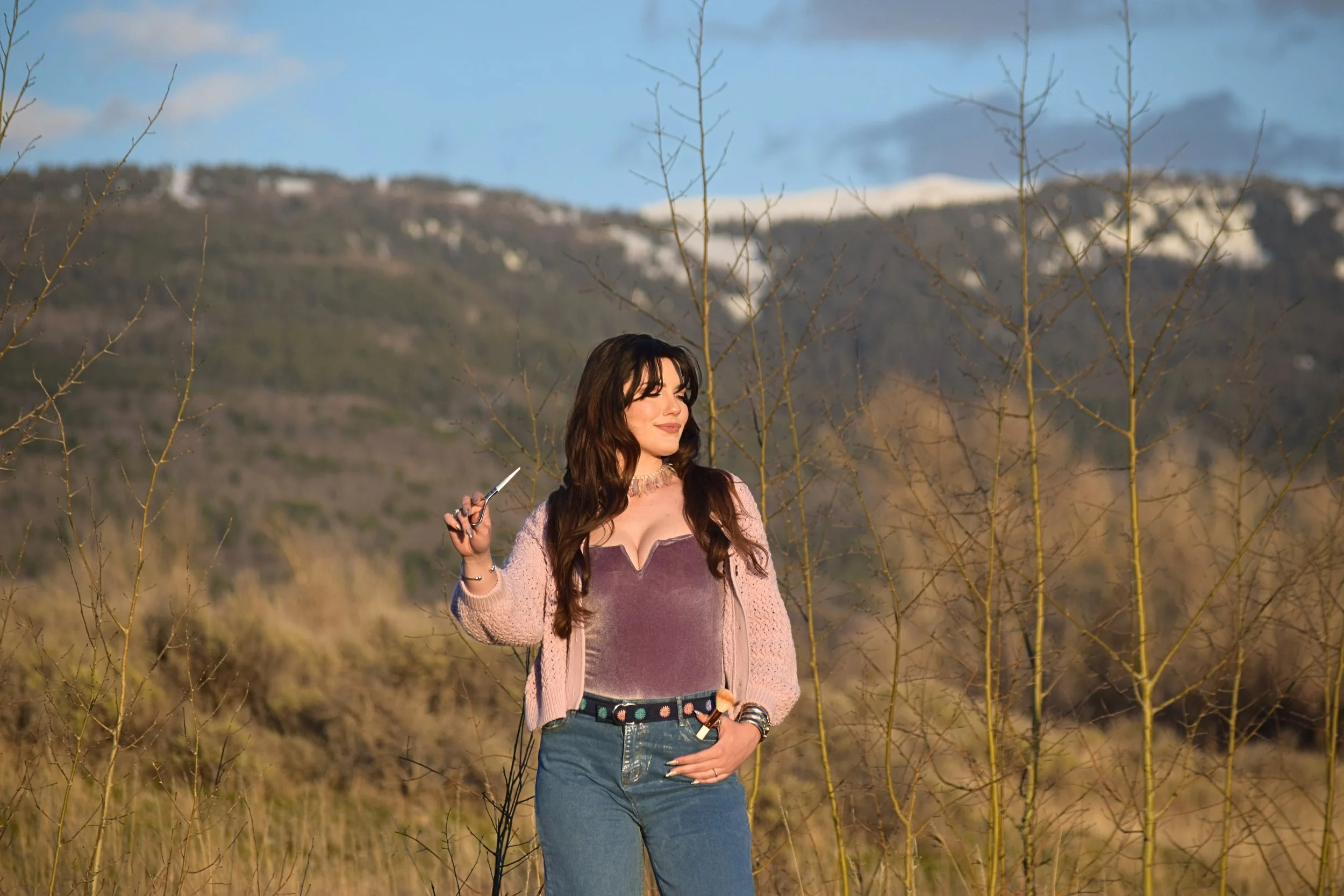 A woman with long brown hair wearing a pink jacket, purple top, and blue jeans stands outdoors in a desert landscape with leafless trees and mountains in the background, holding a knife in her right hand.