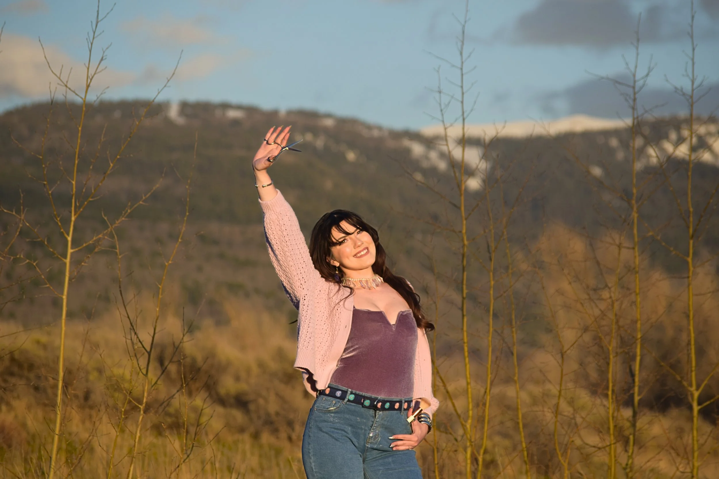 A woman with dark hair, wearing a pink cardigan, a purple velvet top, and blue jeans, standing outdoors in a field with leafless trees and mountains in the background, smiling and waving.