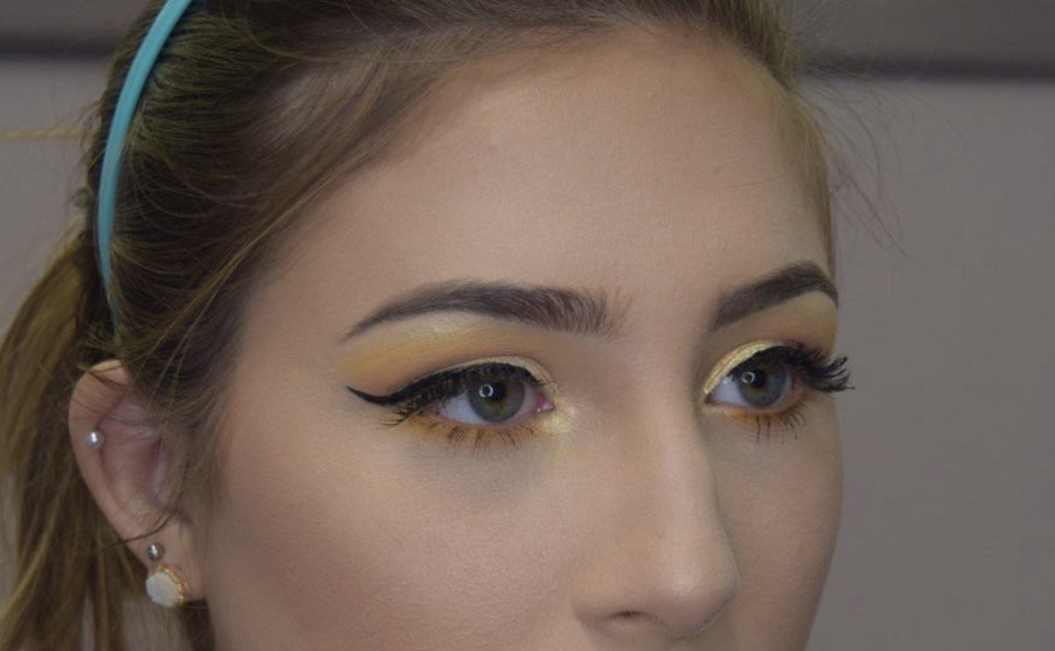Close-up of a woman's face, focusing on her eye makeup with gold eyeshadow and black winged eyeliner, her eyebrows are groomed, and she is wearing earrings with one pearl in each ear, and a blue headband.
