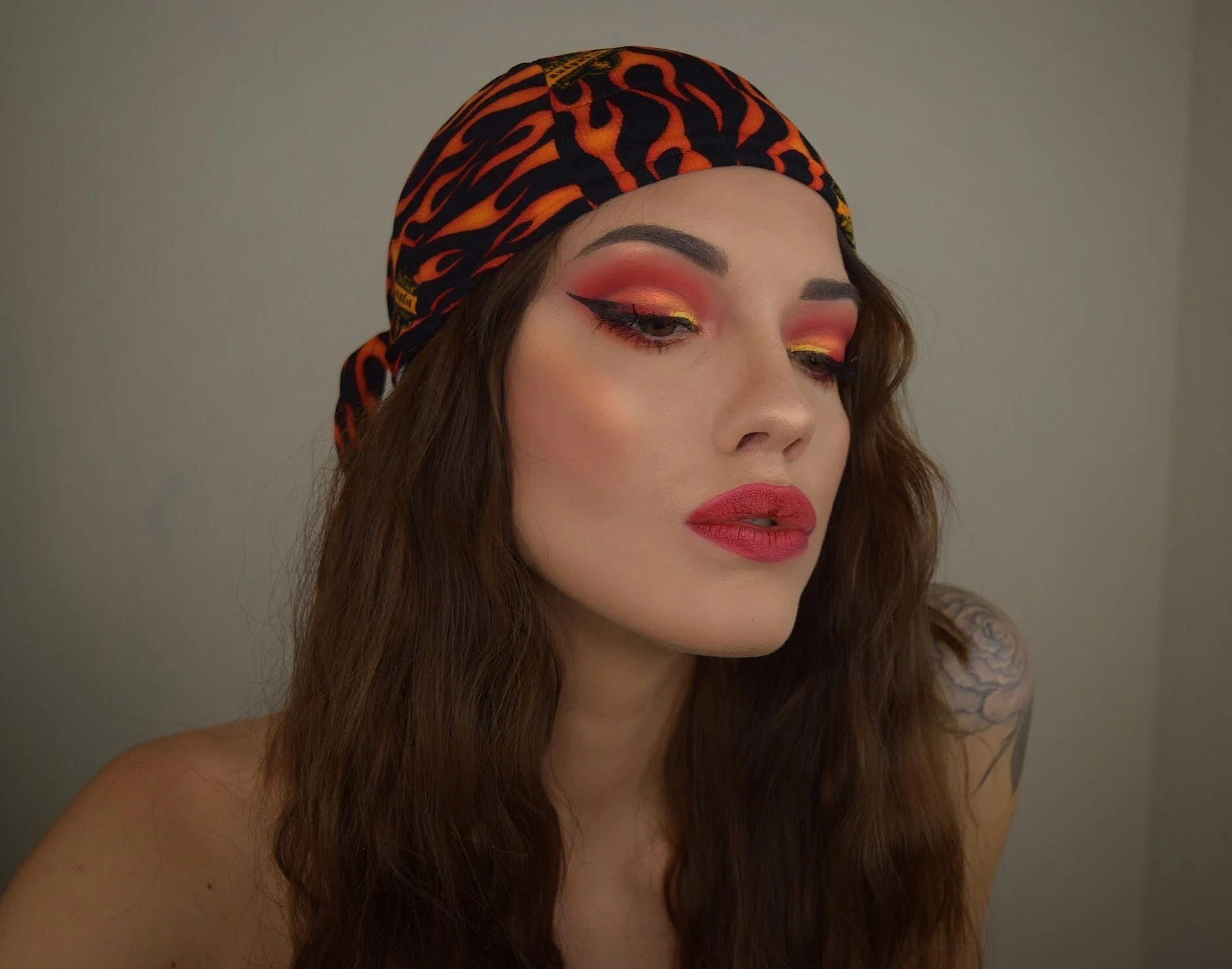 A woman with long brown hair, wearing a black and orange patterned headscarf, has colorful eye makeup with pink, gold, and black eyeliner, and bold red lipstick.