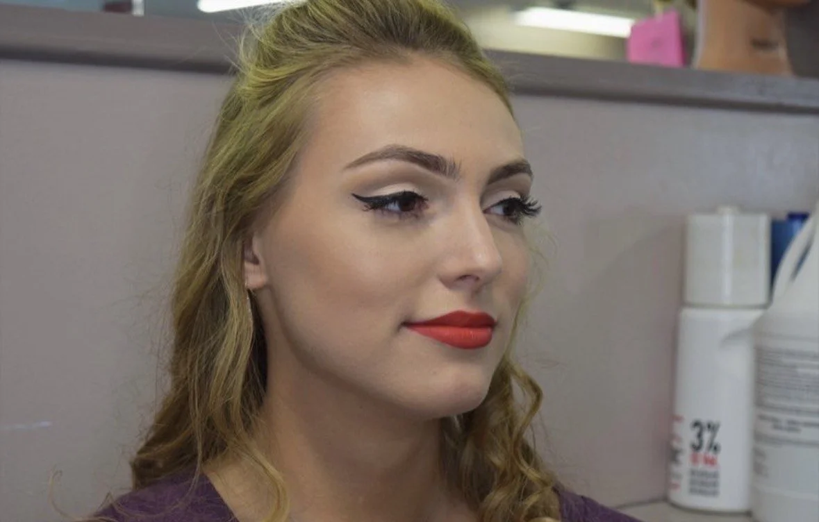 Close-up of a young woman with wavy blonde hair, wearing makeup including winged eyeliner and red lipstick, sitting in an indoor setting with shelves and containers in the background.