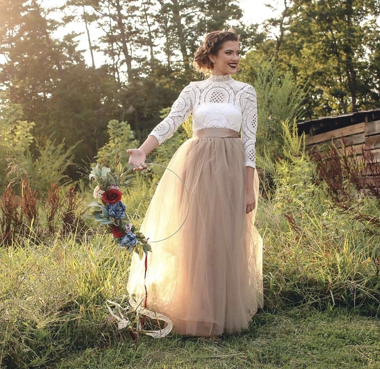 A woman with curly hair in a braided crown, wearing a white lace crop top and a long beige tulle skirt, standing in a grassy field with trees in the background during sunset, holding a floral hoop