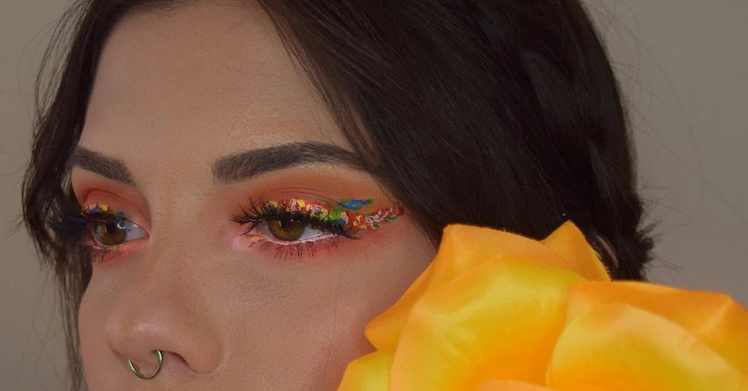 Close-up of a woman with colorful eye makeup and a yellow flower near her face.