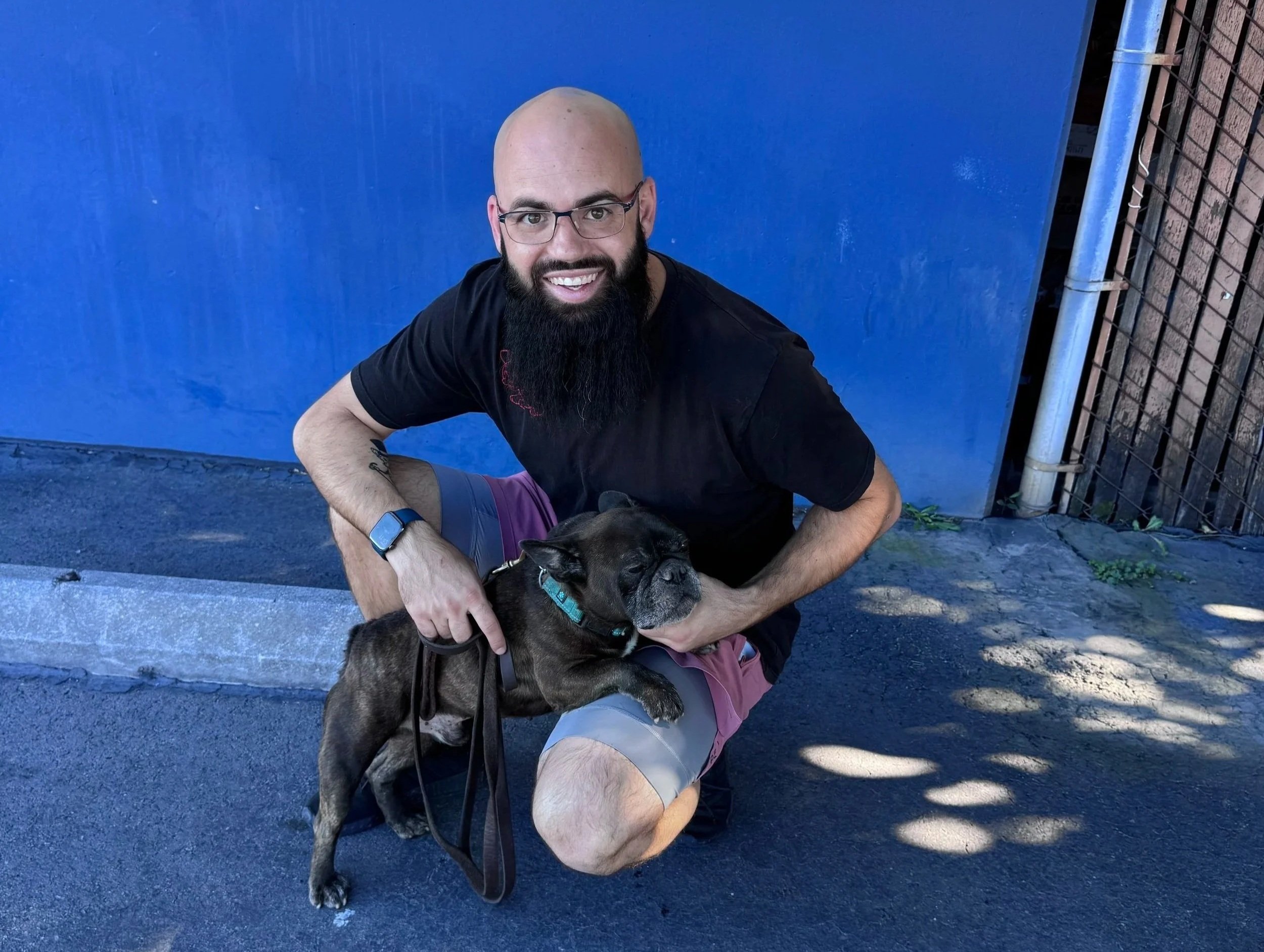 A man with a bald head, glasses, and a beard smiles while crouching next to a brindle French Bulldog with a teal collar. They are outside on a paved area near a blue wall.