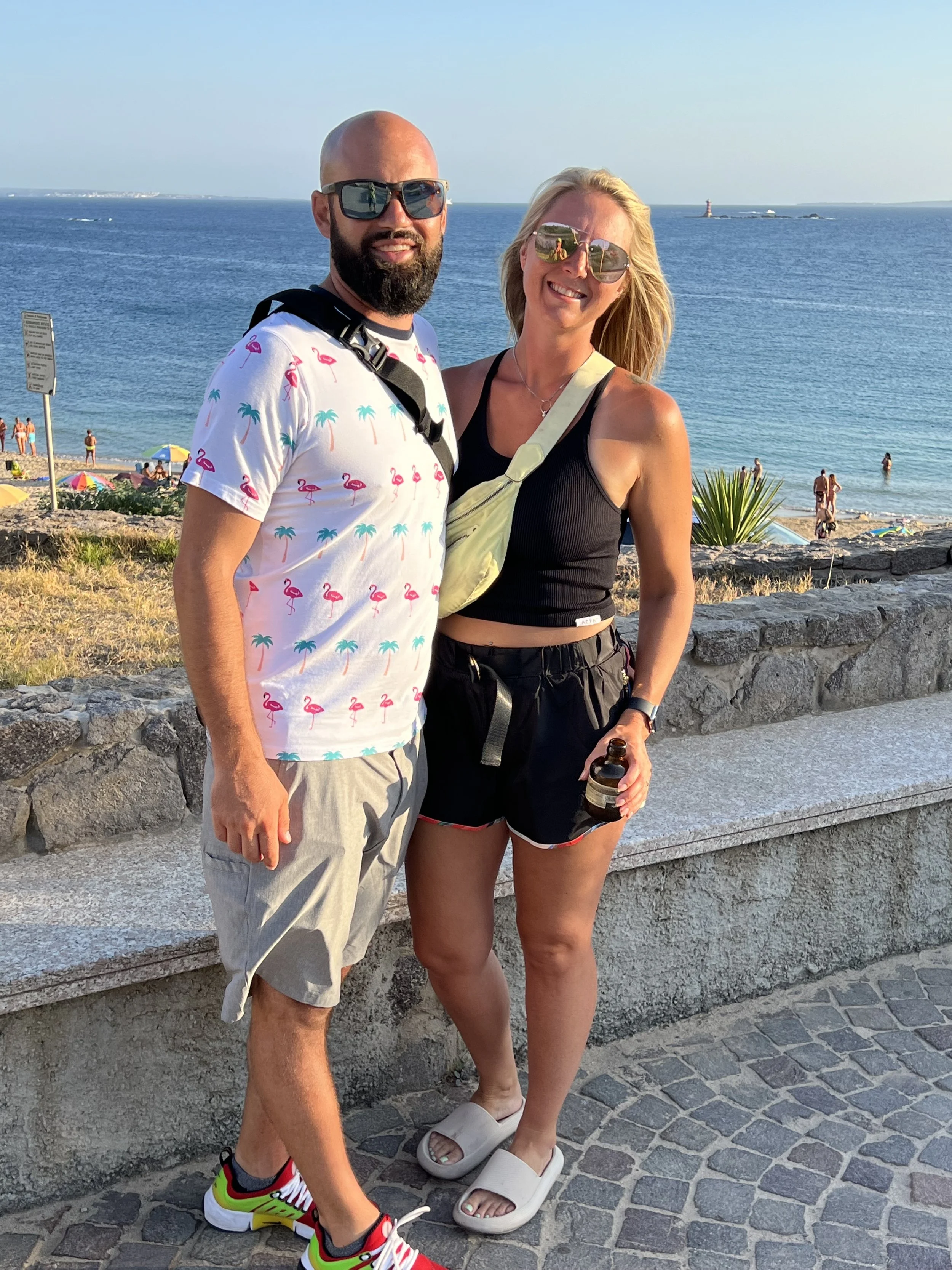 A man and woman standing together on a seaside promenade during sunset, with the ocean and beach in the background, both wearing sunglasses and casual summer clothing.