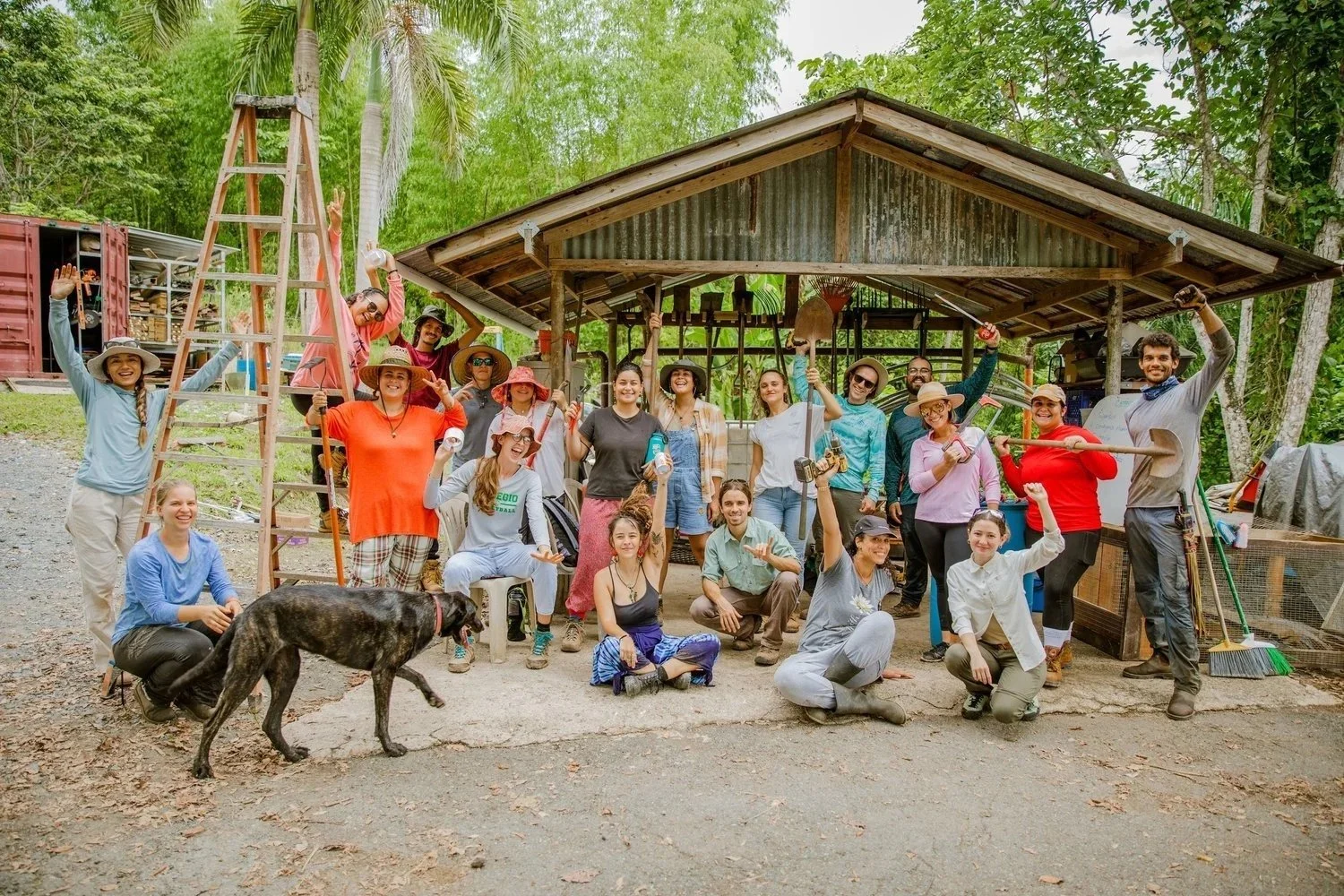 Group of people outdoors holding gardening tools, smiling and celebrating in front of a small shed surrounded by trees.