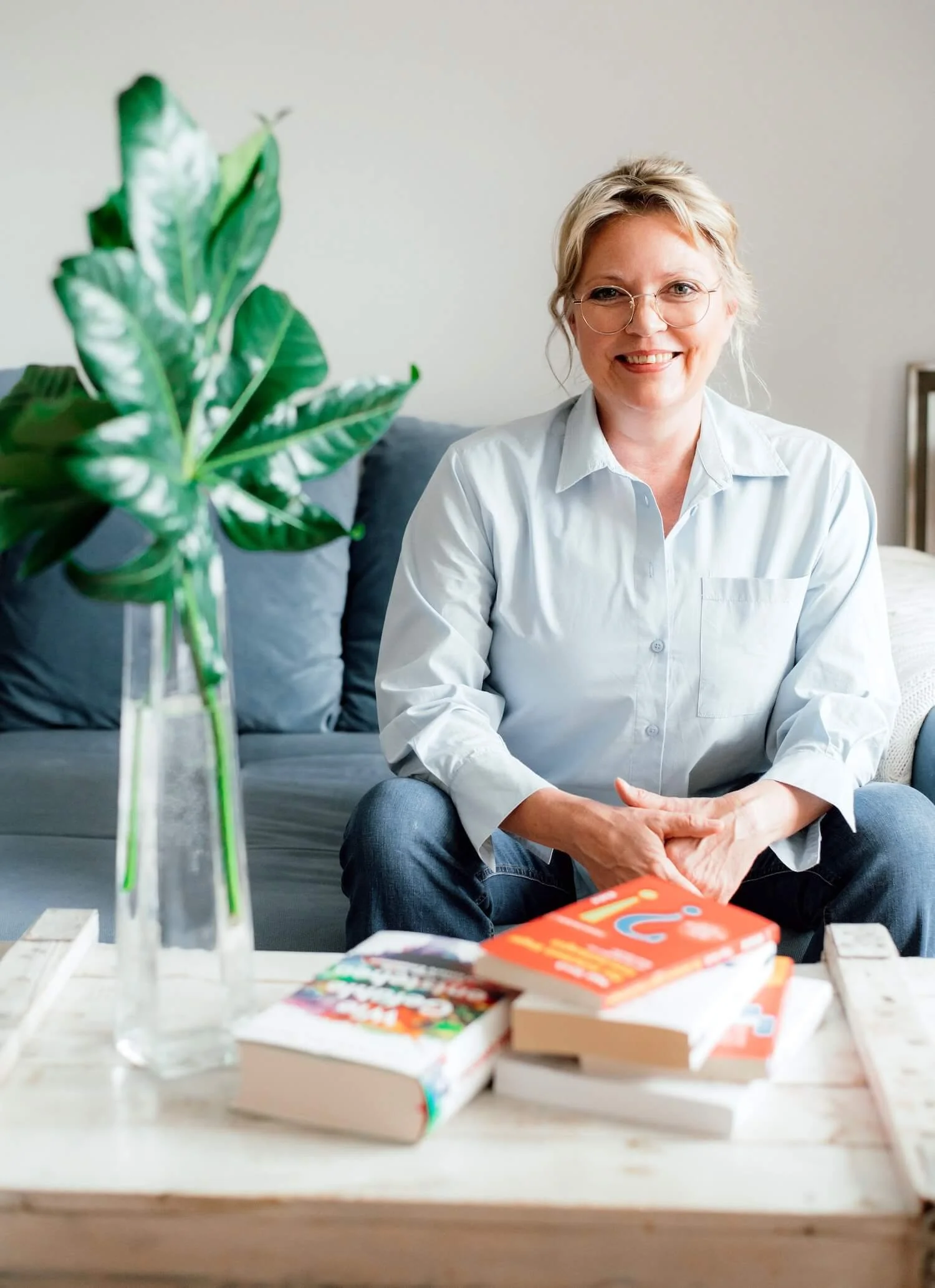 Melanie Friedrich sitzt lächelnd auf einem Sofa und blickt in die Kamera. Im Vordergrund stehen eine grüne Zimmerpflanze in einer Glasvase und Bücher auf einem Couchtisch.