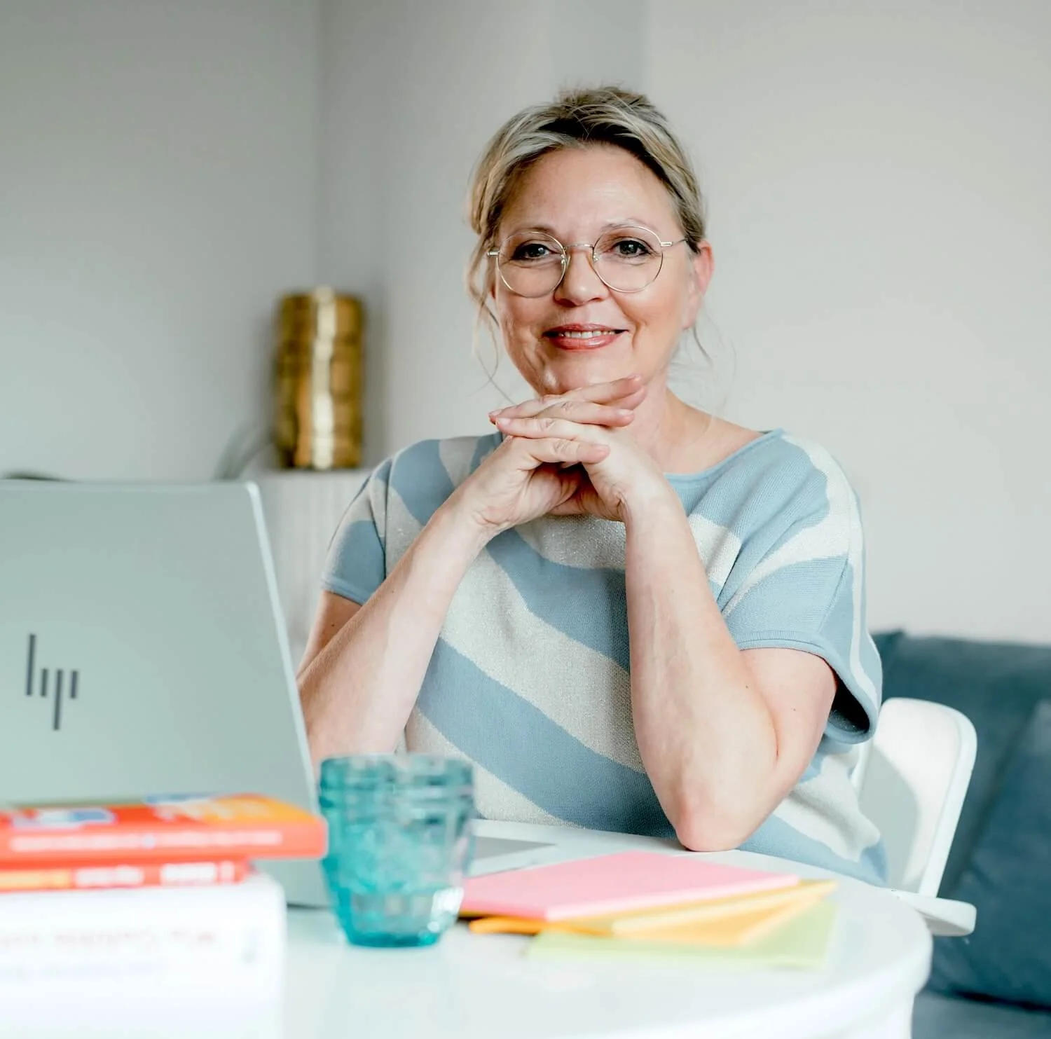 Melanie Friedrich sitzt lächelnd an einem Schreibtisch und blickt in die Kamera. Vor ihr stehen ein Laptop und Bücher, die Hände sind entspannt aufeinandergelegt.