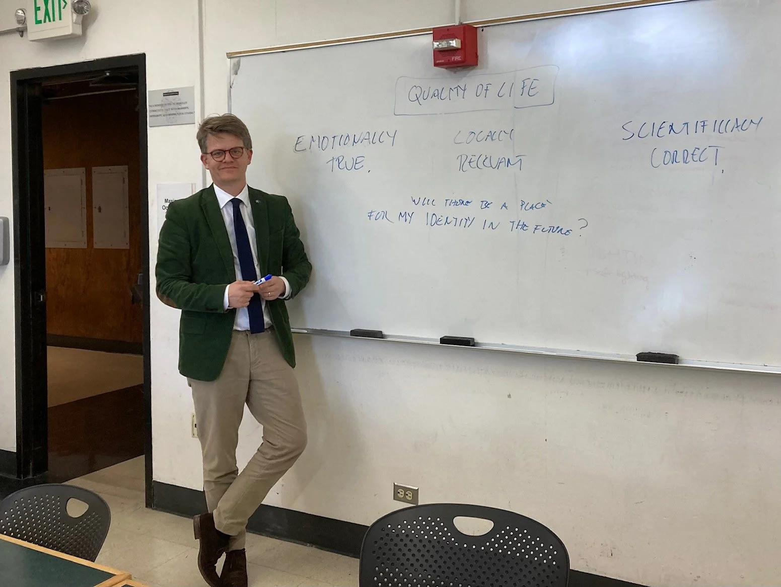 A man with glasses and a green blazer standing next to a whiteboard with handwritten text, in a classroom or conference room.