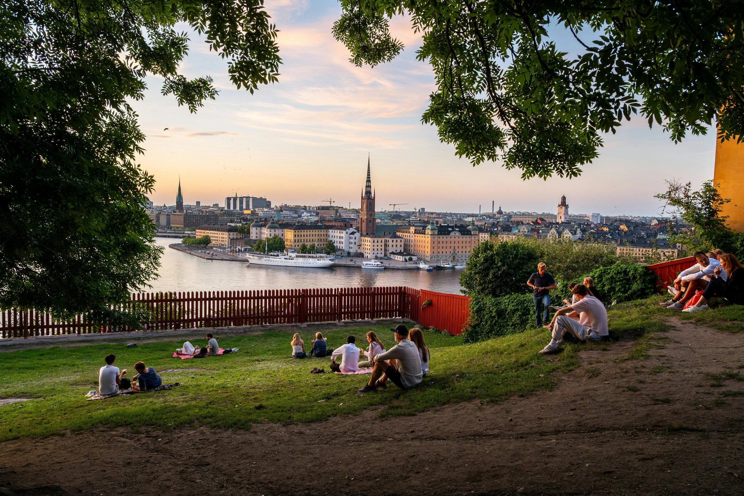 People relaxing on a grassy hillside overlooking a river and a city skyline during sunset, with trees framing the scene.