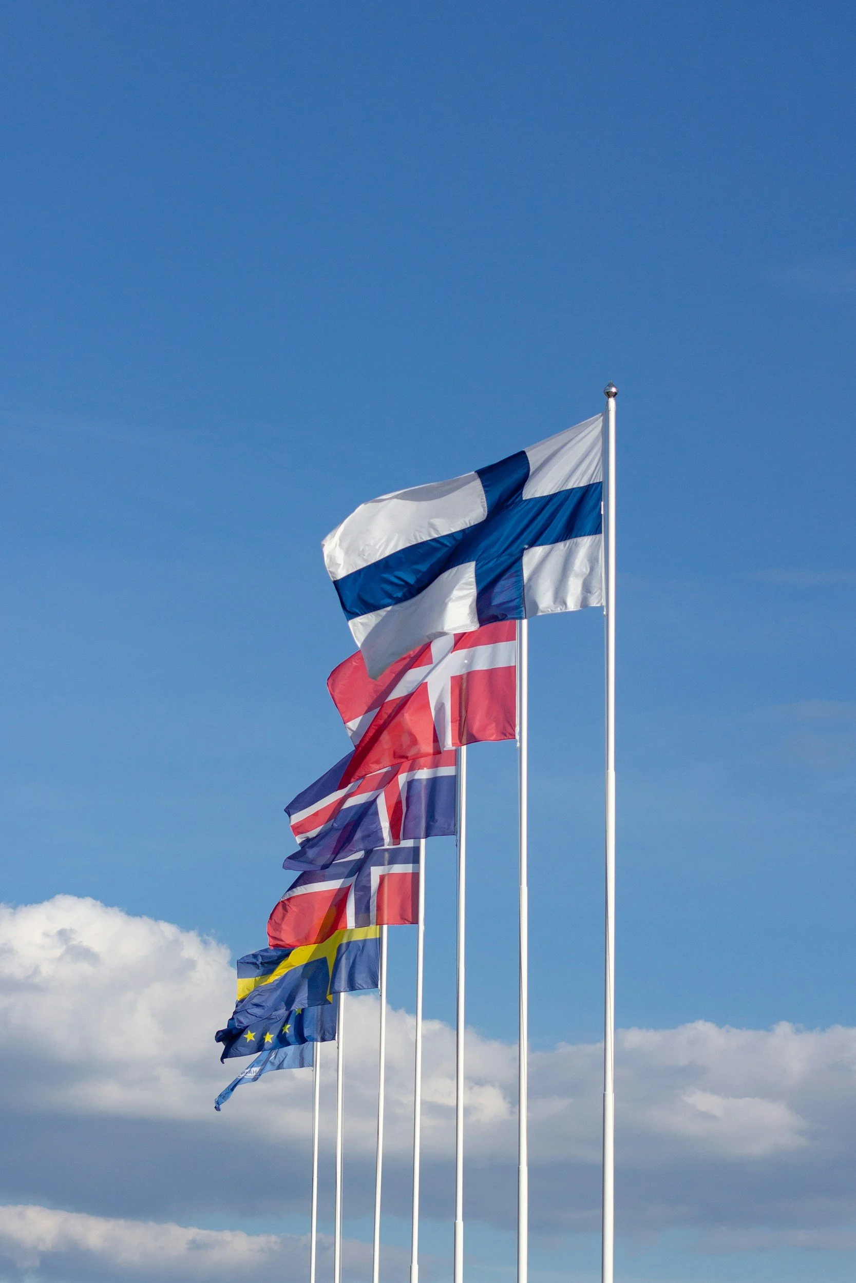 Multiple flags, including the Finnish flag, flying on flagpoles against a blue sky with some clouds.
