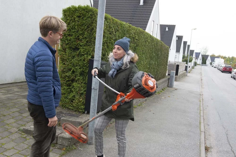 A woman with gray hair and a winter coat holds a string trimmer while talking to a man in a blue jacket on a sidewalk in a residential neighborhood.