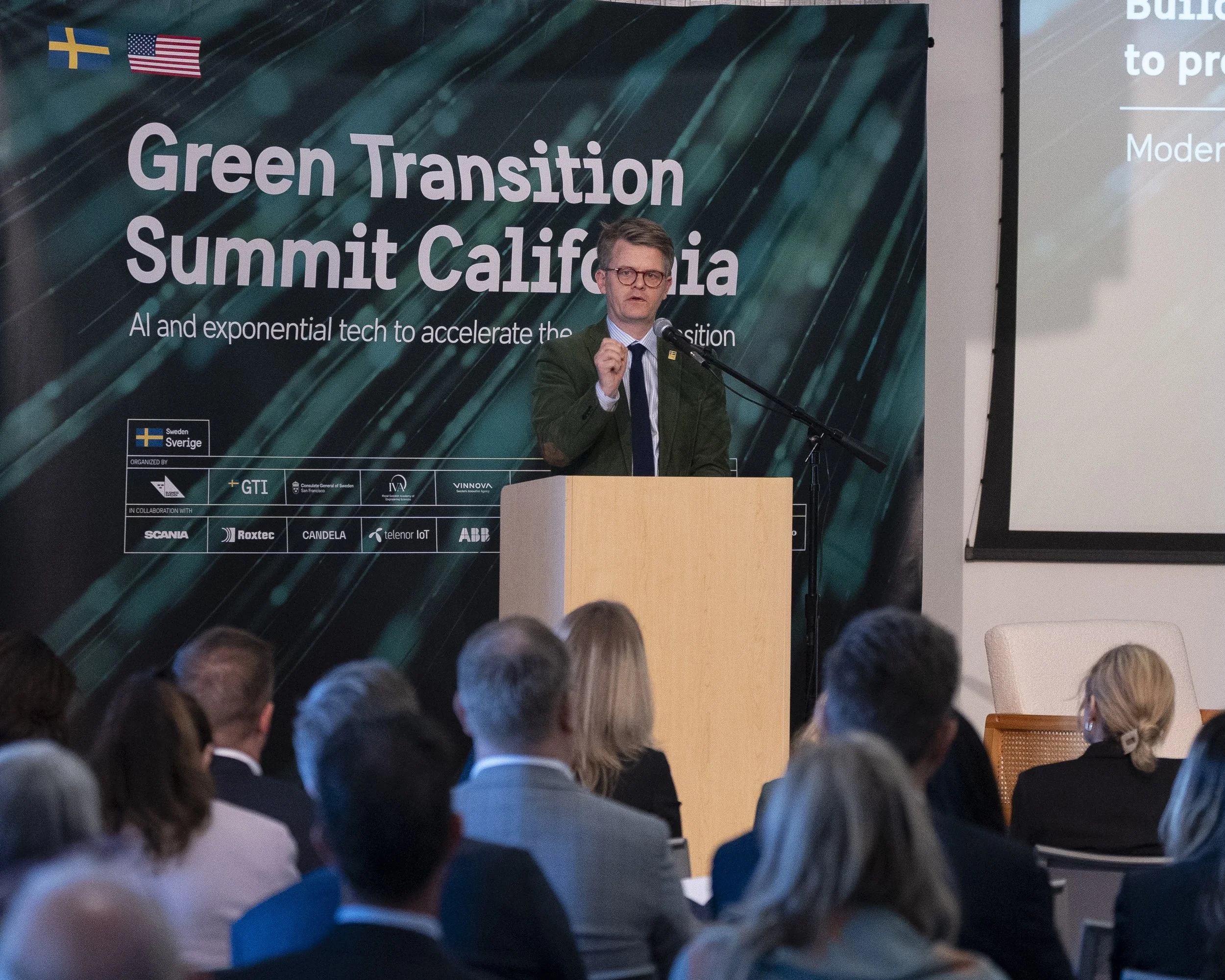 A man in a green jacket and glasses is speaking at a podium during the Green Transition Summit California, with an audience and a large banner in the background. The banner displays flags of Sweden and the United States and sponsors' logos.