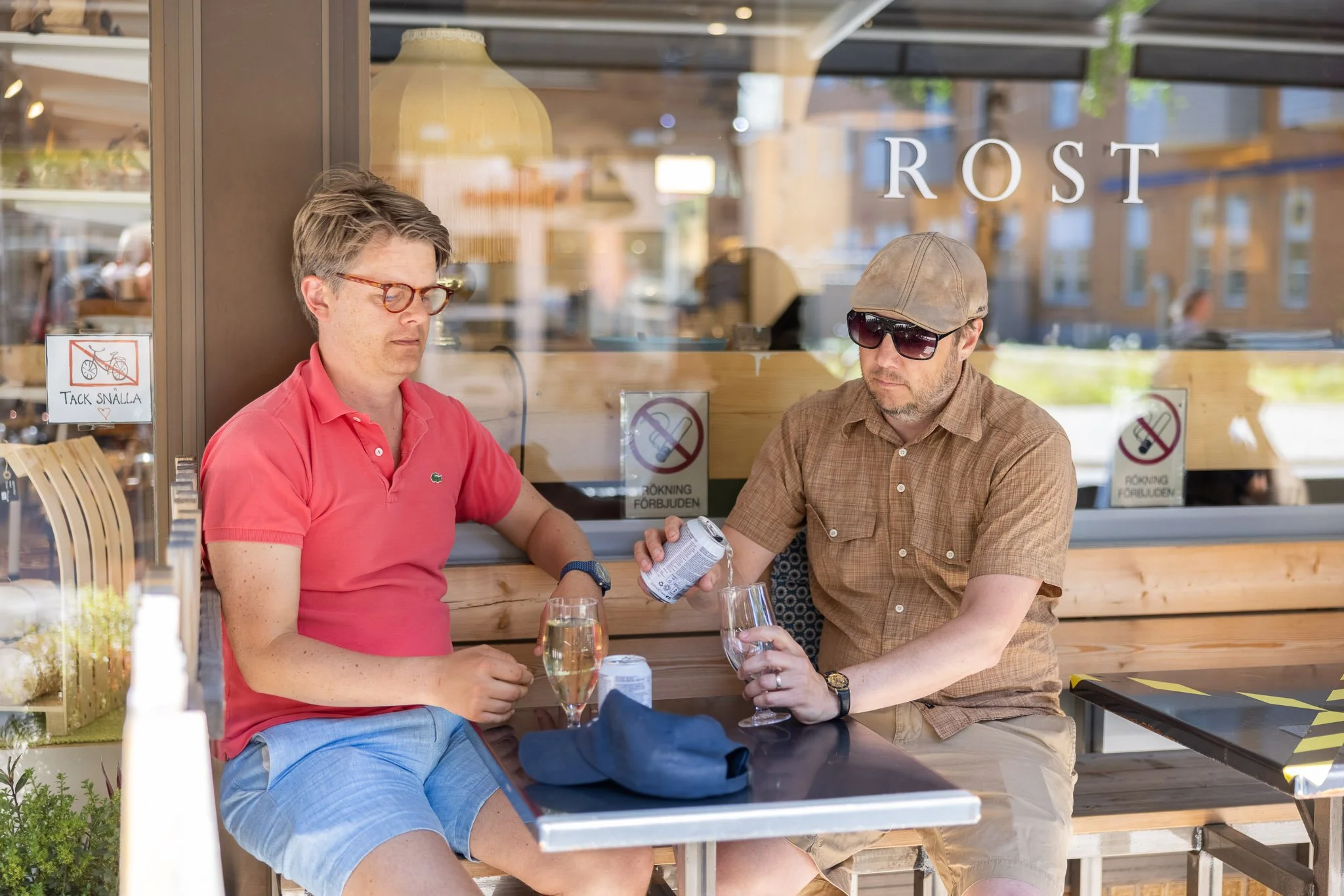 Two men sitting outside at a cafe table, one pouring a drink into a glass, with signs in Swedish on the window behind them.