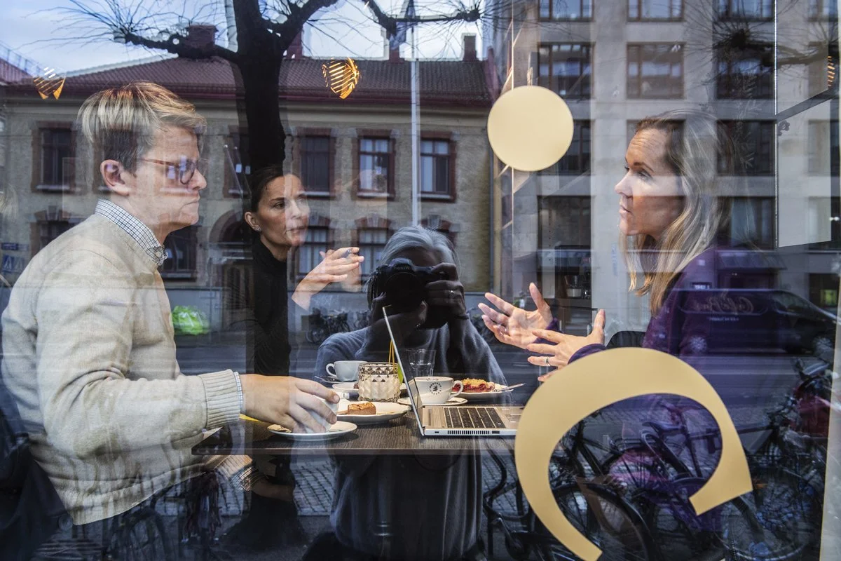 A group of four people having a discussion inside a cafe, seen through the large window, with reflections of the street and buildings outside.