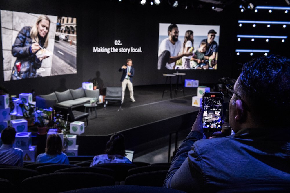 A man taking a photo with his phone of a speaker on stage at a conference, with two large screens behind him showing different images and text about making stories local.