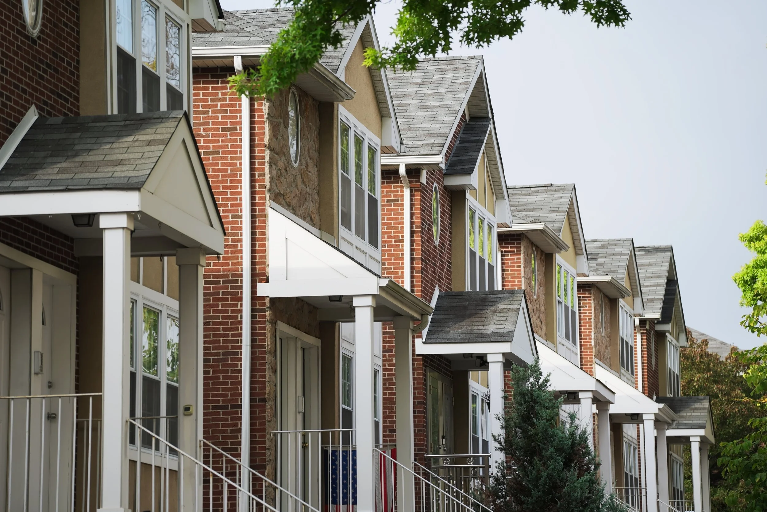 Row of townhouses with brick and stucco exteriors, gable roofs, and front porches with white railings and stairs, surrounded by green trees.
