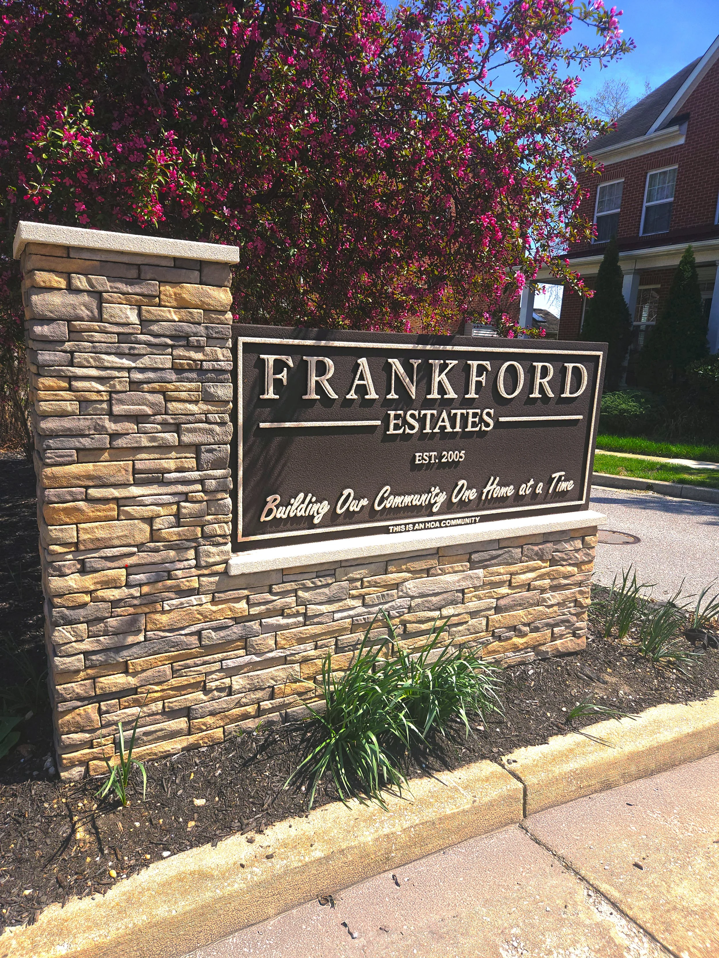 A stone sign that reads "Frankford Estates" with the slogan "Building Our Community One Home at a Time," located near a blooming pink flowering tree and a brick house in the background.