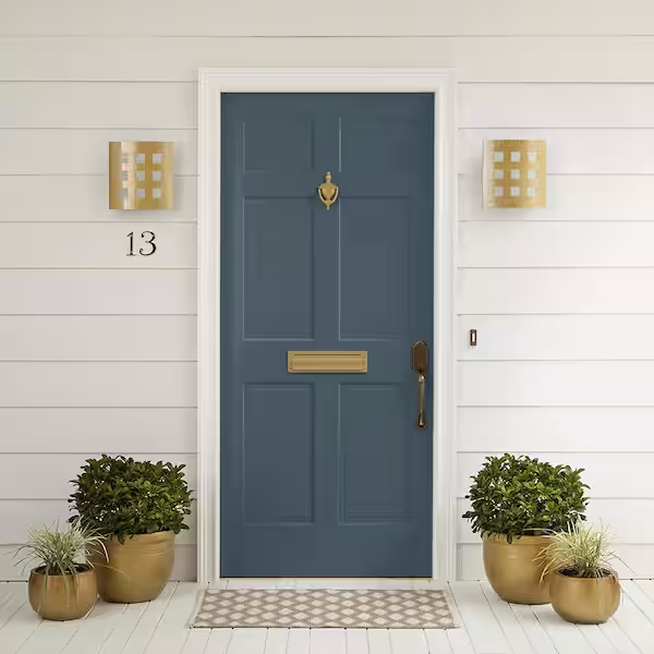 Front door with house number 13, blue door with gold handle and mail slot, white siding, two gold wall lights, two potted bushes, and a small rug.