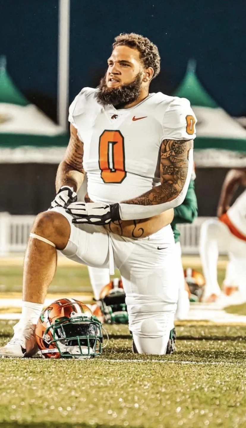 A football player with a beard and tattoos, wearing a white jersey with the number 0, kneeling on one knee on the field at night. His helmet is off and placed on the ground in front of him. He appears to be looking ahead with a serious expression.