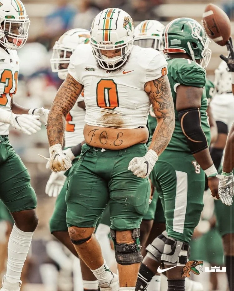 A football player from the Miami Hurricanes wearing a white jersey with the number 0 and green pants, with tattoos on his arms and stomach that reads 'F.O.E.' I see other players in green jerseys around him.