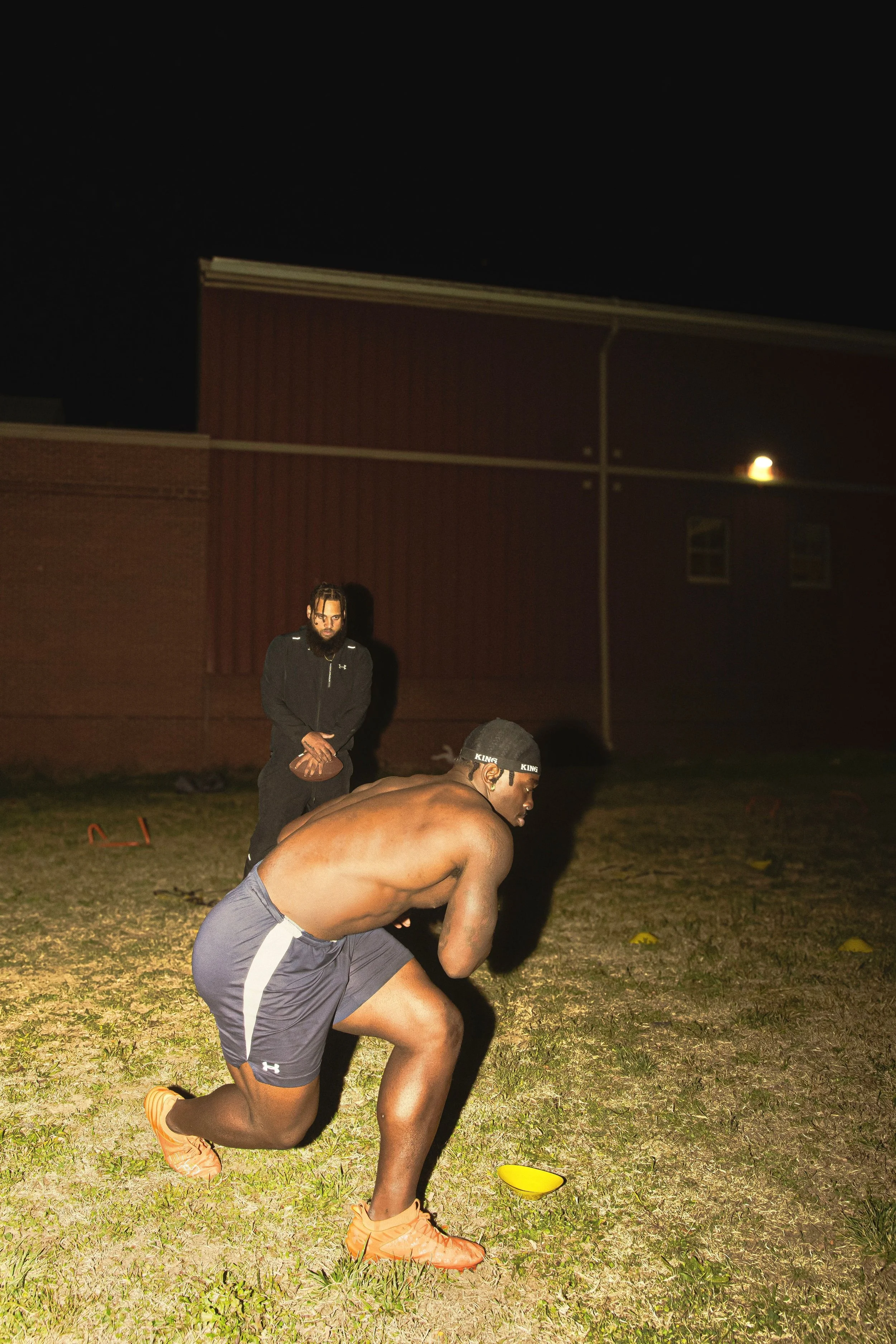 A shirtless man in athletic shorts and orange cleats is kneeling on the grass in a football stance during nighttime practice, with a coach or trainer standing behind him holding a football.
