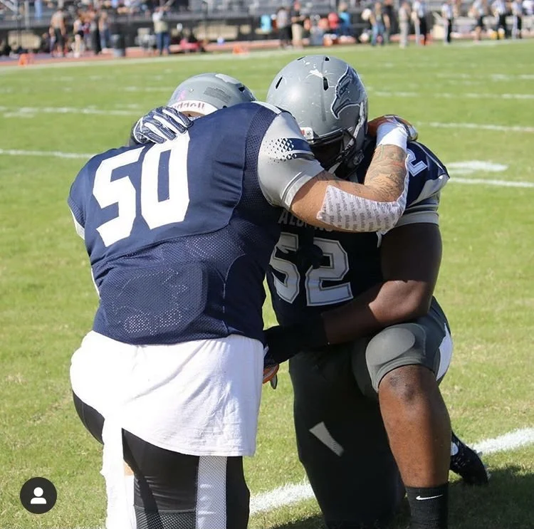 Two football players from the same team wearing helmets and jerseys are hugging on the field.